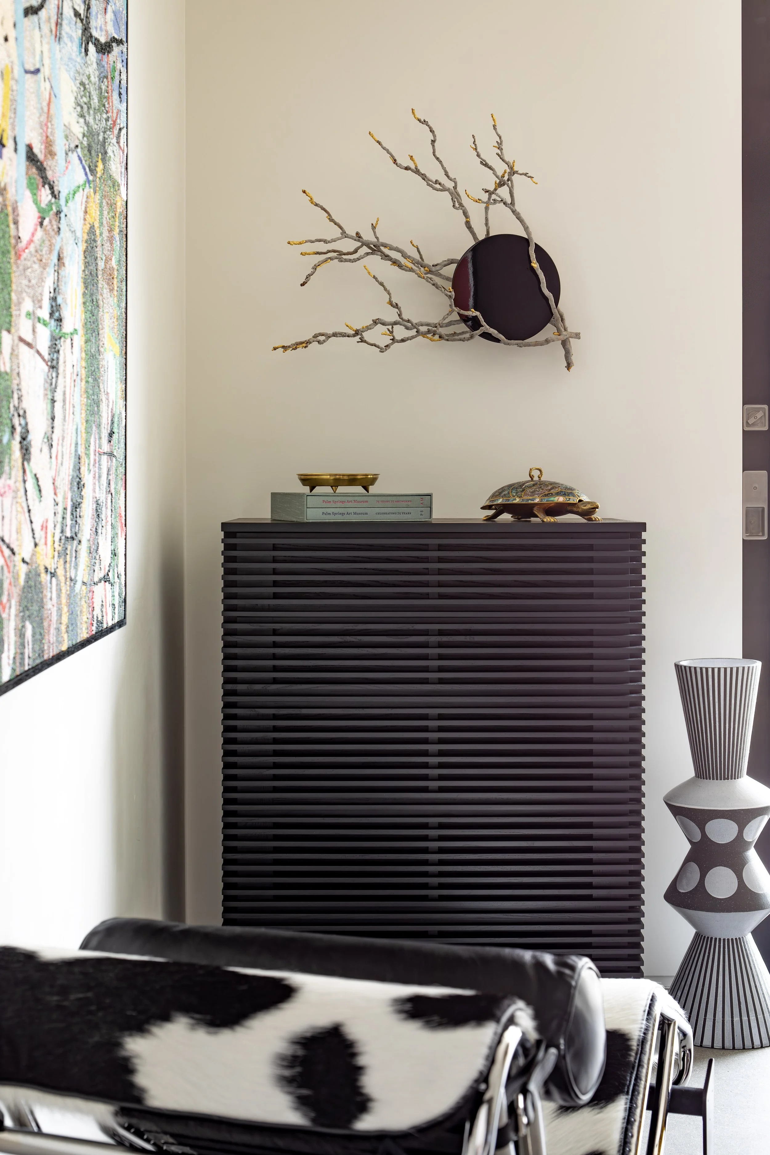 Modern interior corner with a black slatted cabinet, decorative objects on top, a branch wall sculpture, a patterned ceramic vase, and a colorful framed artwork on the wall.