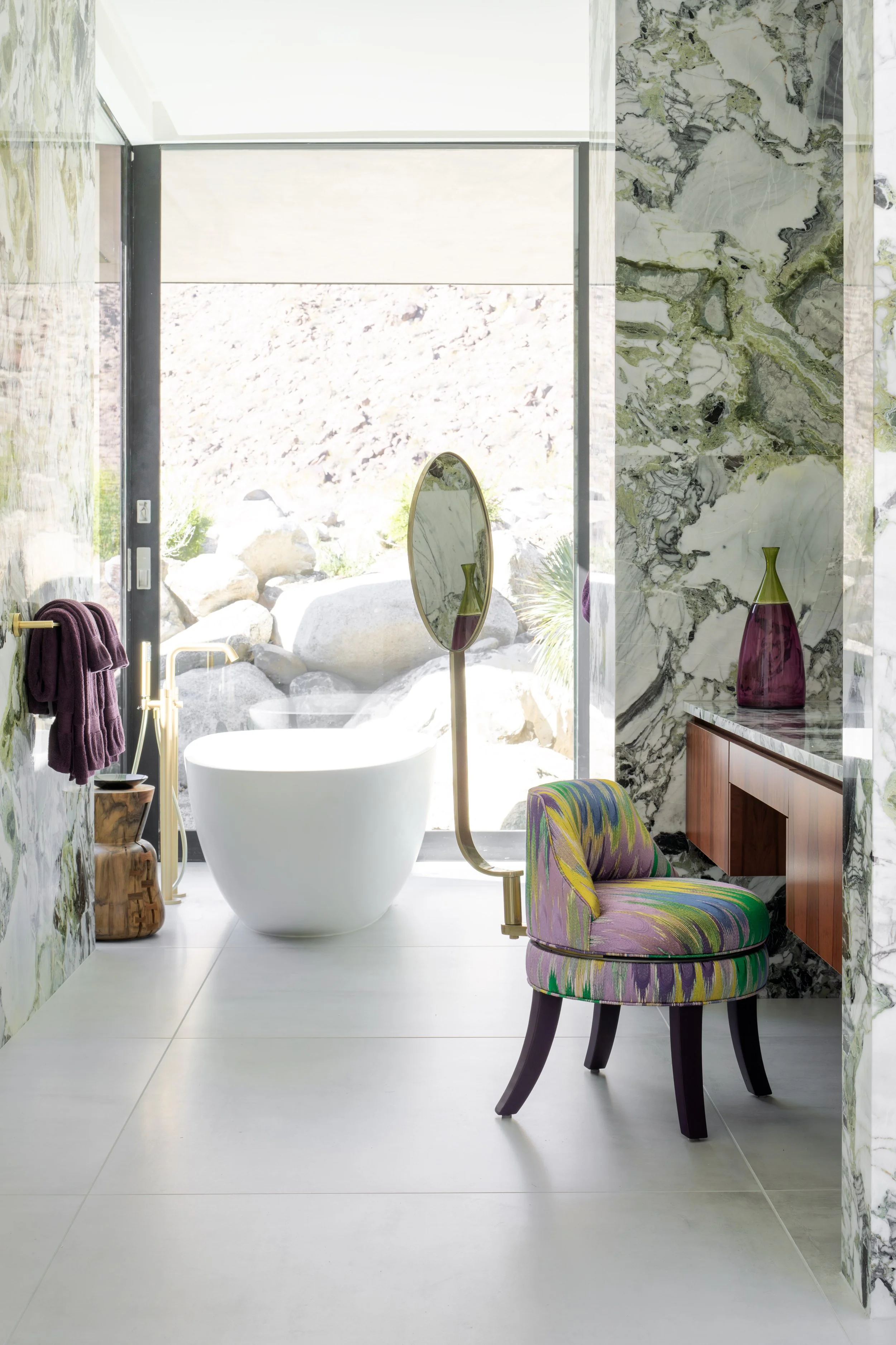 Modern bathroom with a freestanding bathtub near large glass sliding doors, marble walls, colorful upholstered chair, and decorative vases.