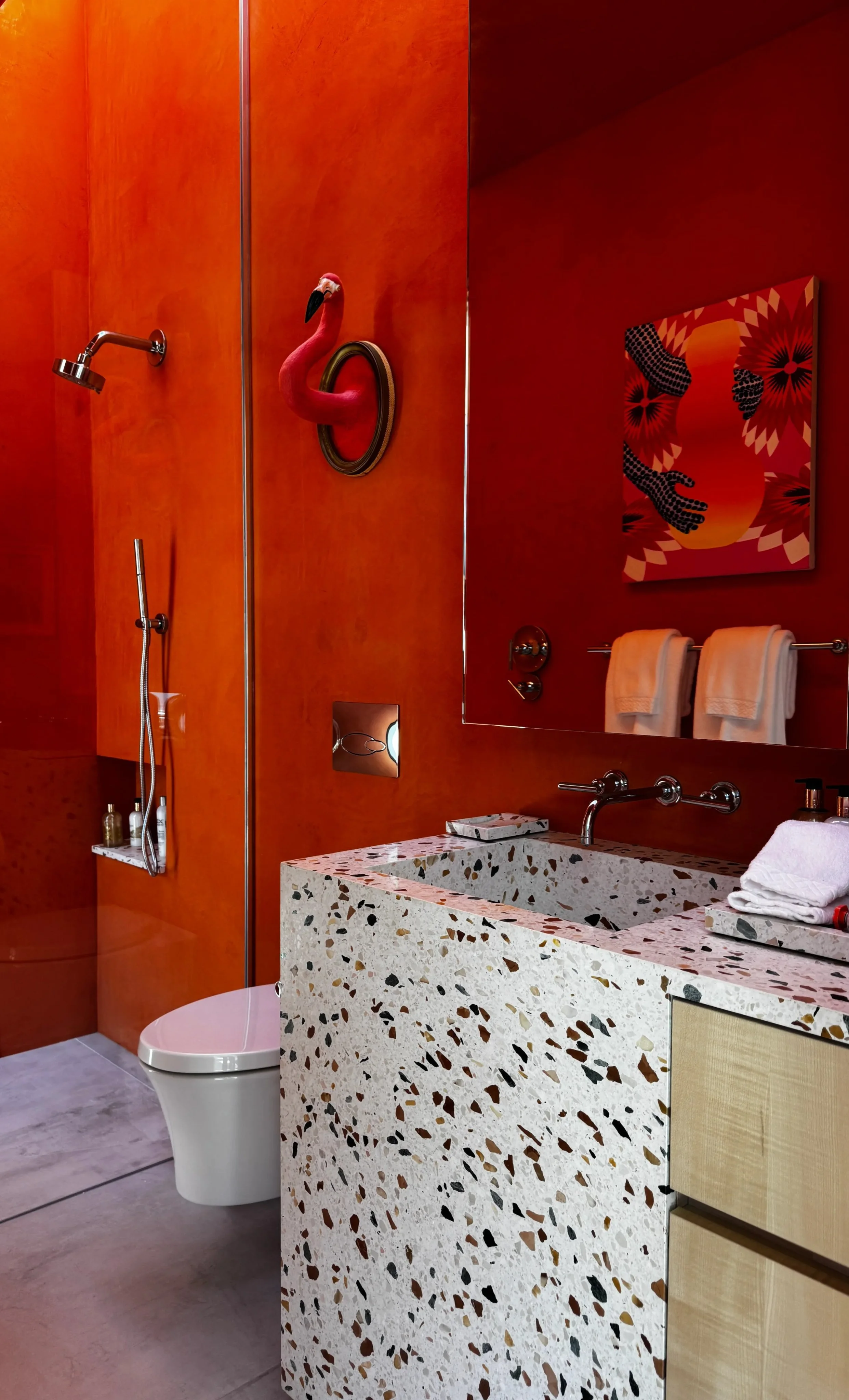 A bathroom with burnt orange walls, a terrazzo sink, a large mirror, a walk-in shower with a mounted showerhead, towel racks with white towels, and colorful artwork of a flamingo and abstract art on the wall.