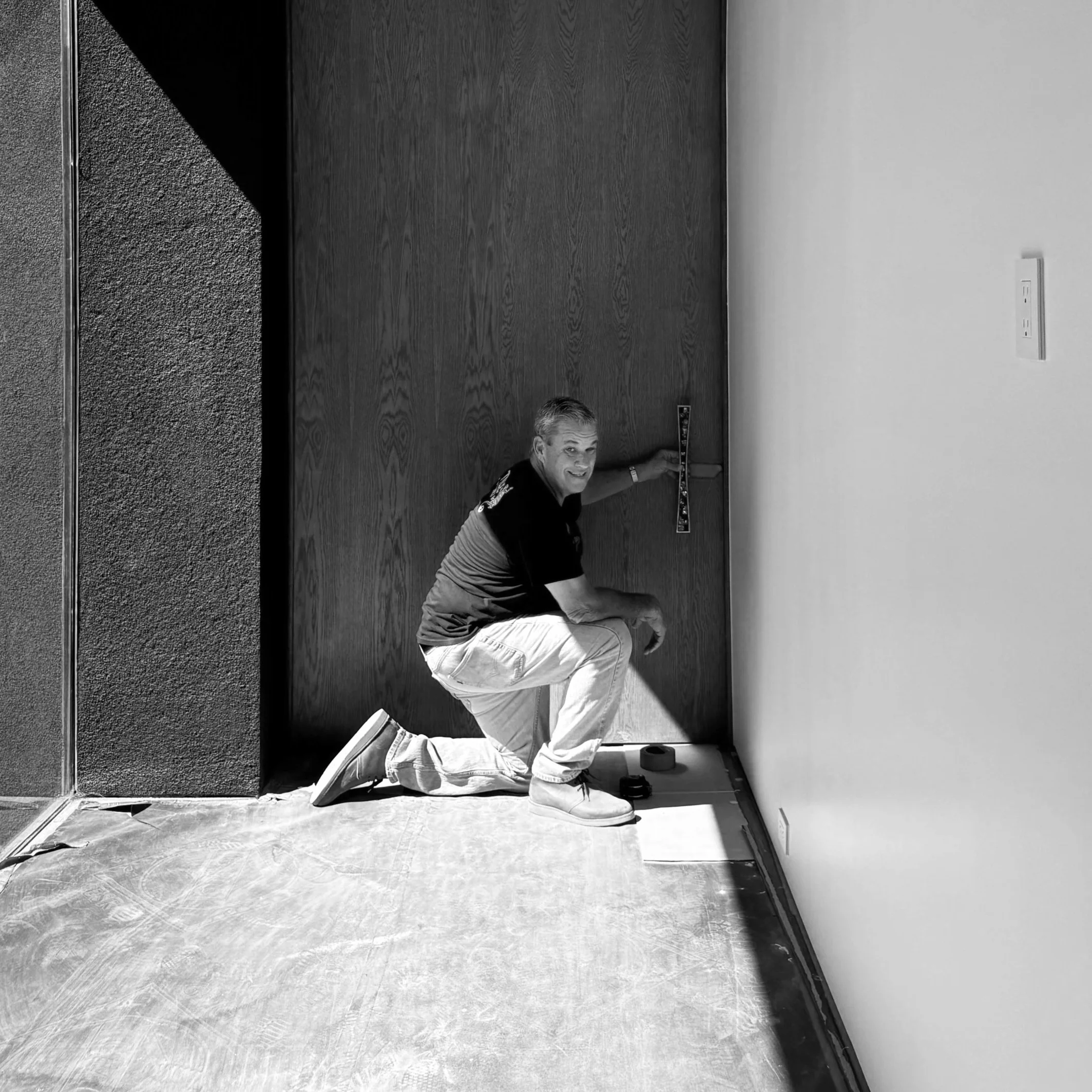 A man kneeling on the floor installs or measures a door frame in a room, holding a level against the door. The room has one textured wall, a plain wall with a light switch, and a window on the left side.