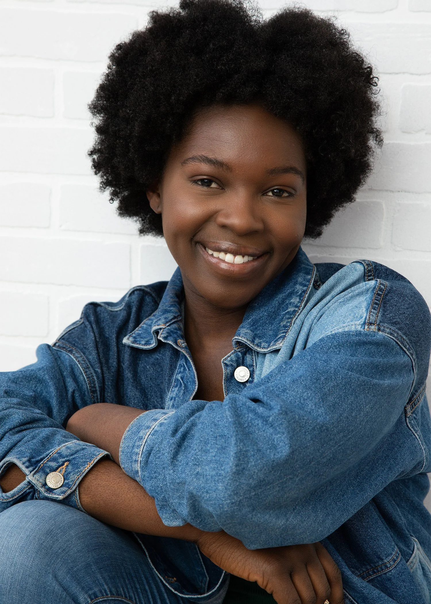 Portrait of a smiling woman with short curly black hair, wearing a denim jacket, sitting against a white brick wall.
