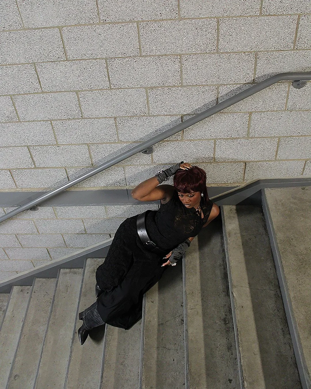 A woman on a staircase looking up at the camera, resting her hand on her head, dressed in black with jewelry and gloves.