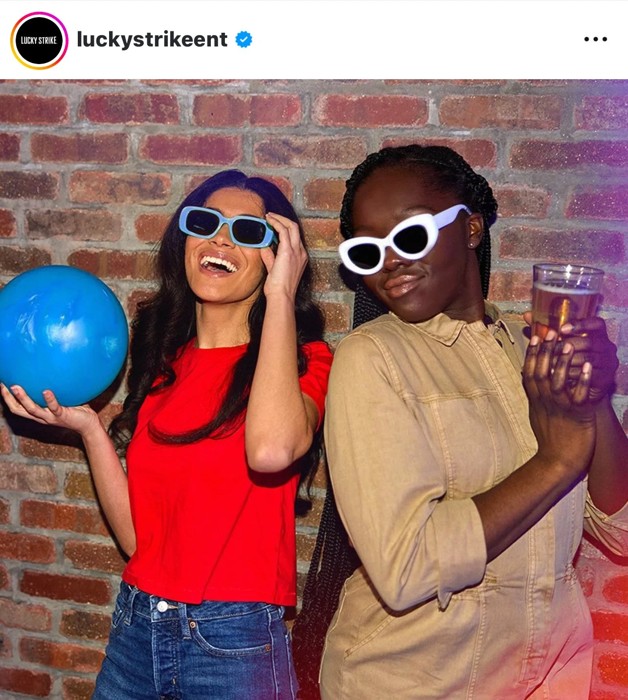 Two women wearing sunglasses, one with blue glasses holding a blue ball and the other with white glasses holding a glass of clear beverage, standing against a brick wall, smiling and having fun at a party or celebration.