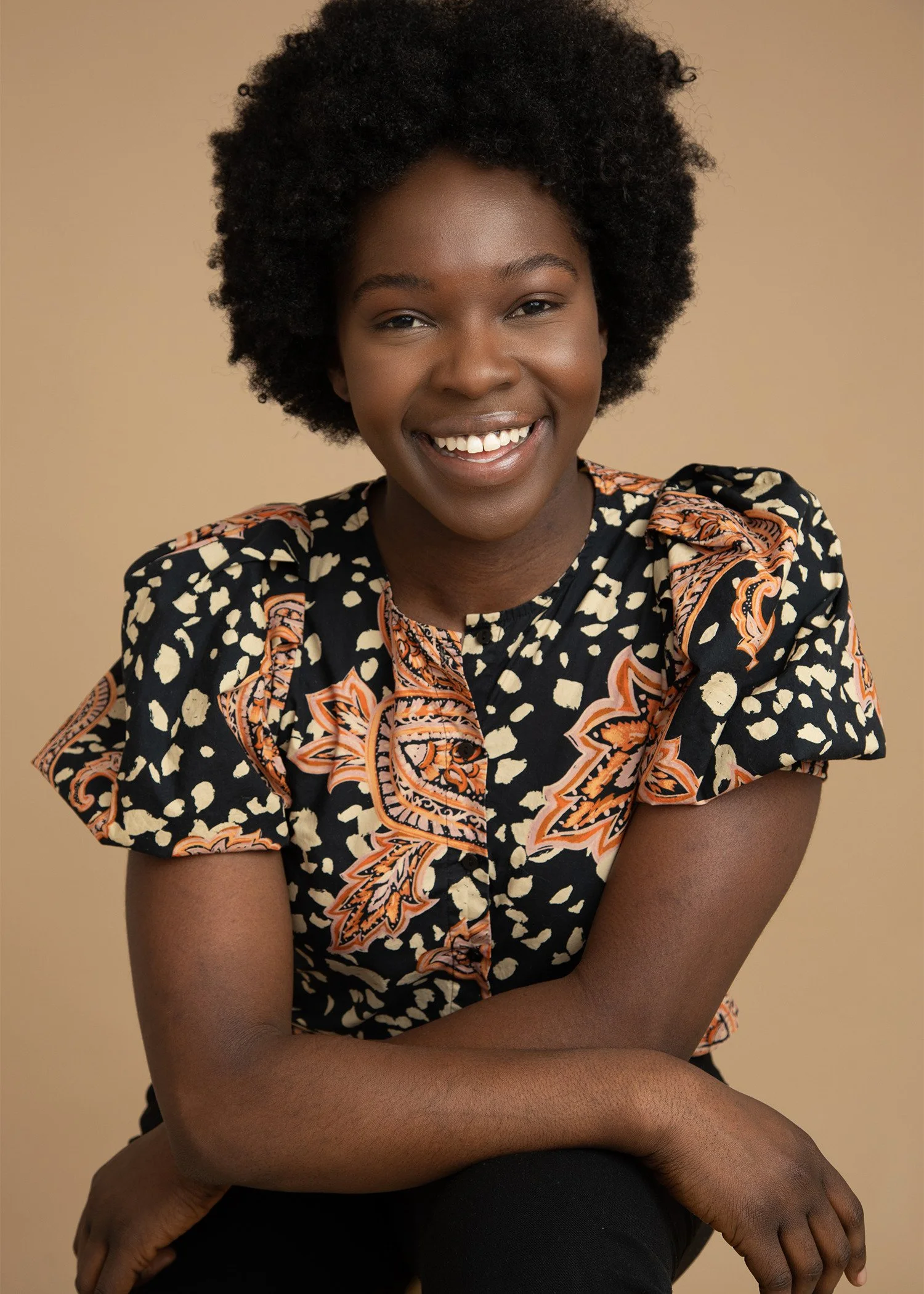 A young woman with natural curly hair, smiling, wearing a patterned black, orange, and cream blouse, sitting against a beige background.