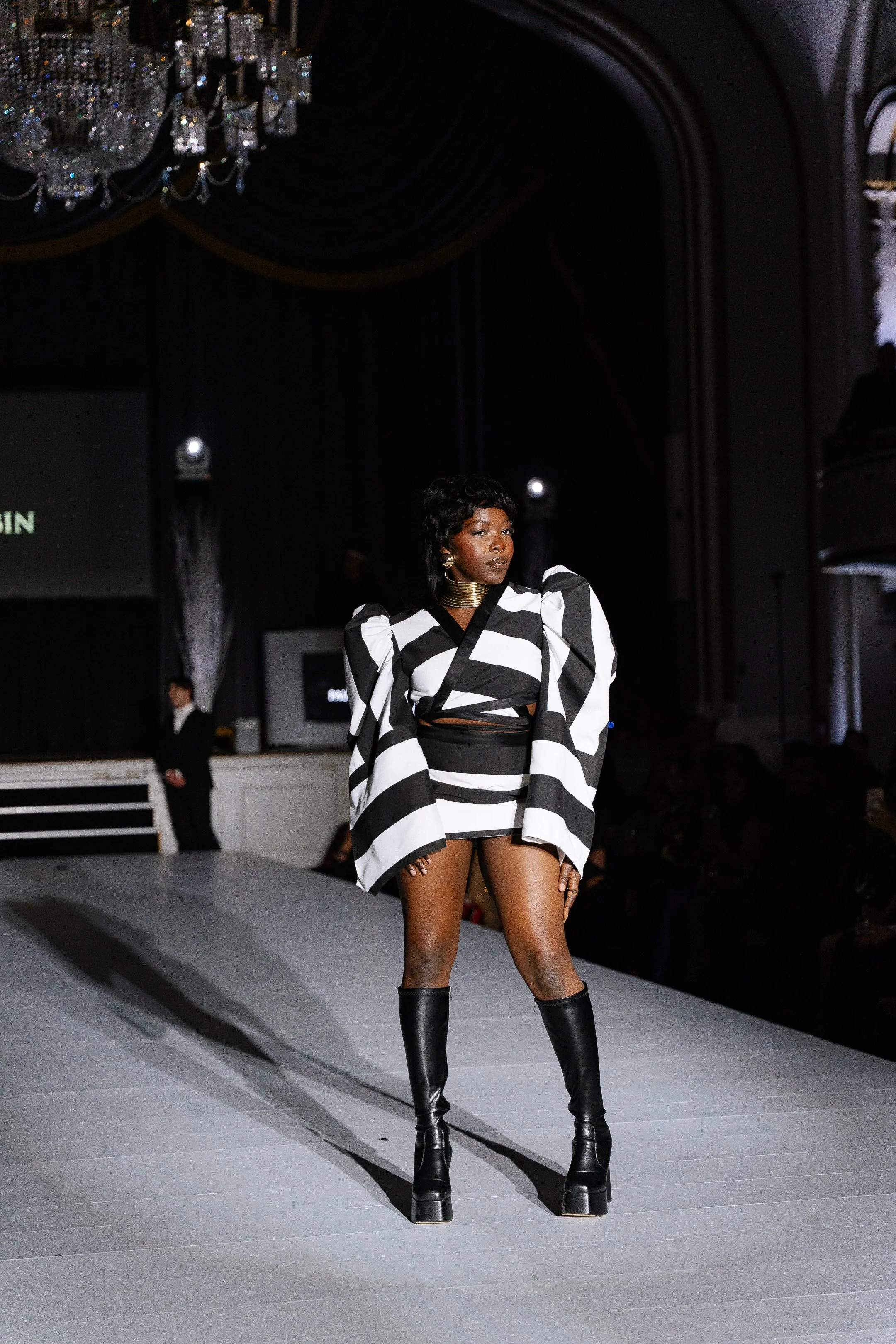 Model wearing a black and white striped outfit with puffed sleeves and black knee-high boots walking on a runway at a fashion show with a chandelier overhead.
