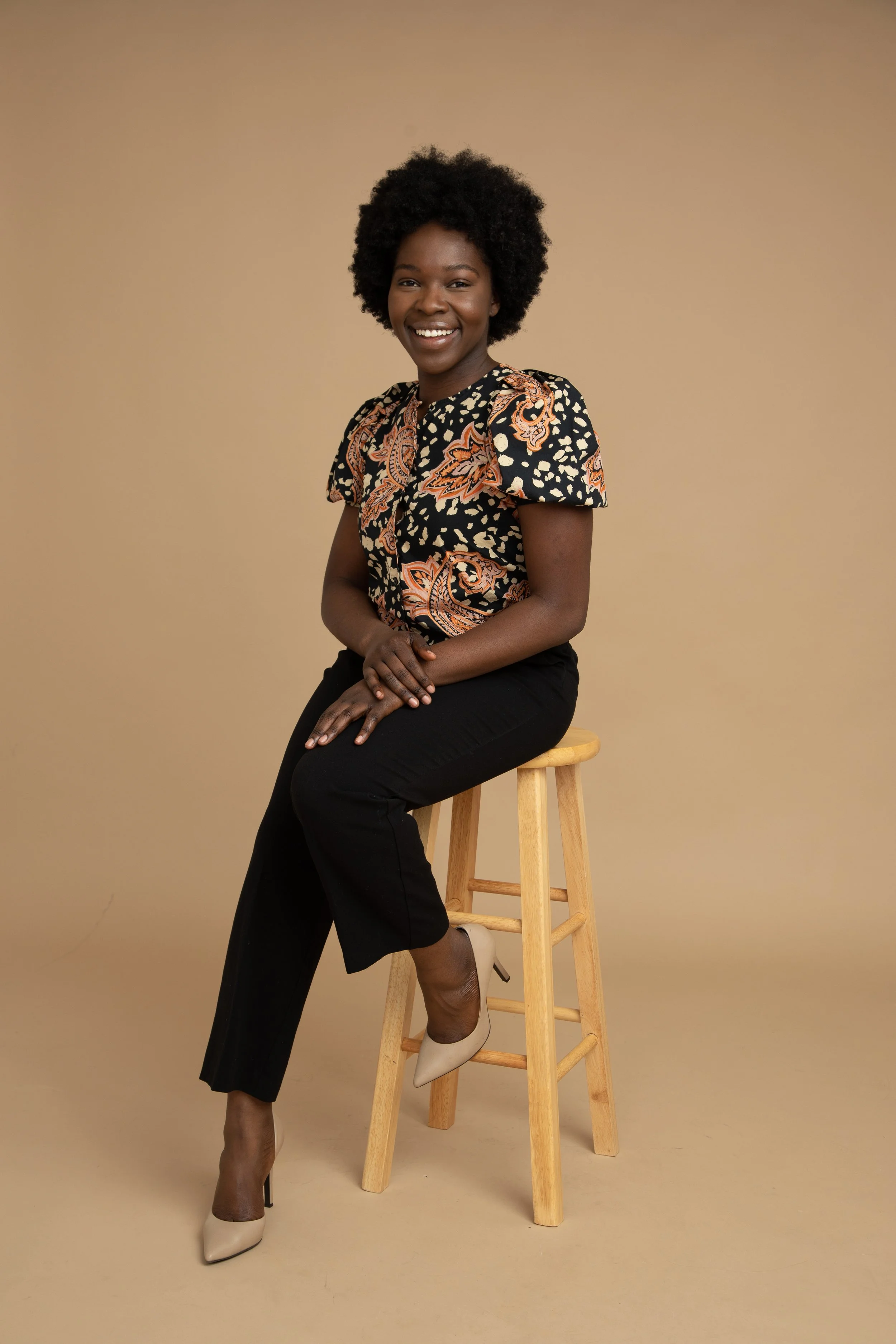 A woman sitting on a wooden stool, smiling, wearing a patterned blouse, black pants, and beige high heels, against a beige background.