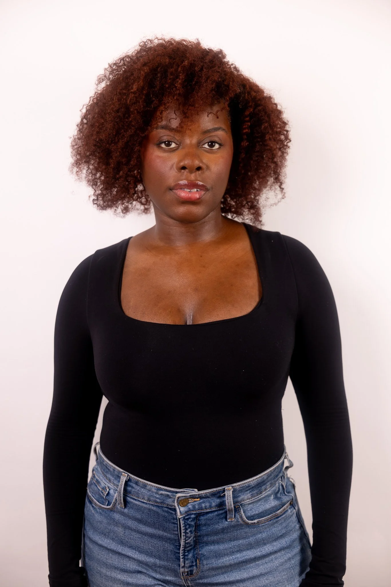 Portrait of a Black woman with curly reddish-brown hair wearing a black long-sleeve top and blue jeans against a plain white background.