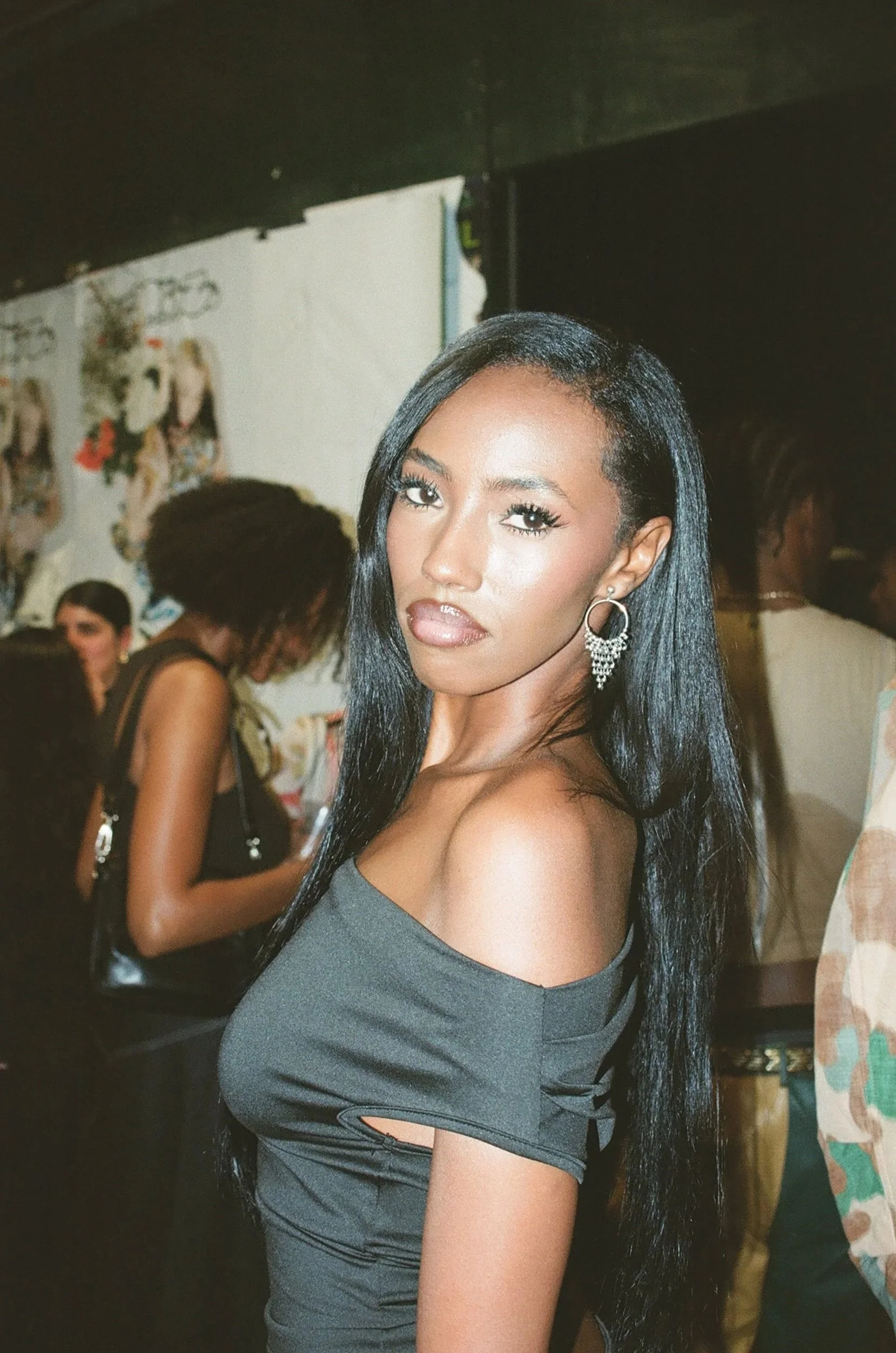 A woman with long black hair, wearing a dark off-shoulder dress and large earrings, looking at the camera at an indoor event with several other people in the background.