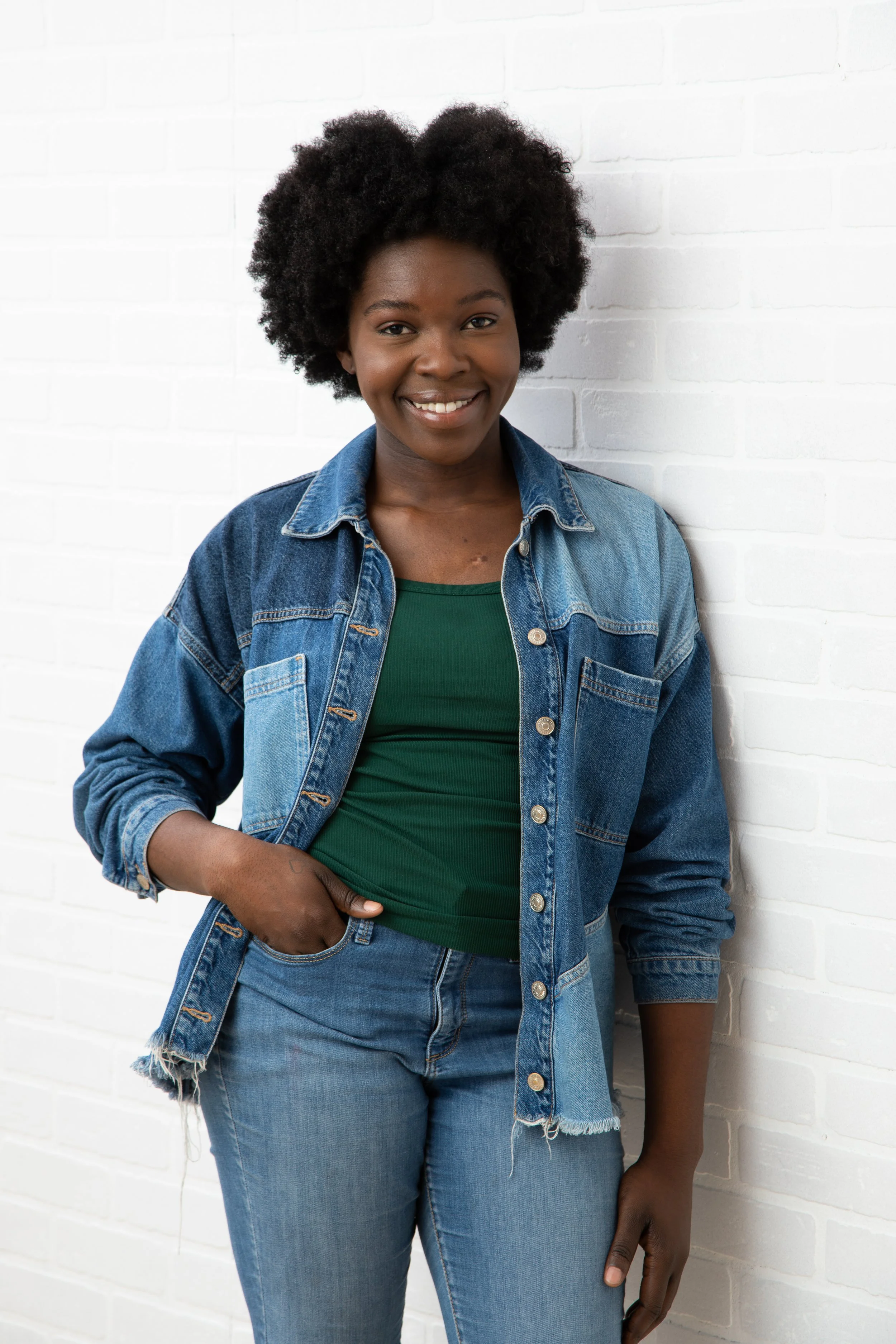 A smiling woman with curly black hair, wearing a green shirt, denim jacket, and jeans, stands against a white brick wall.