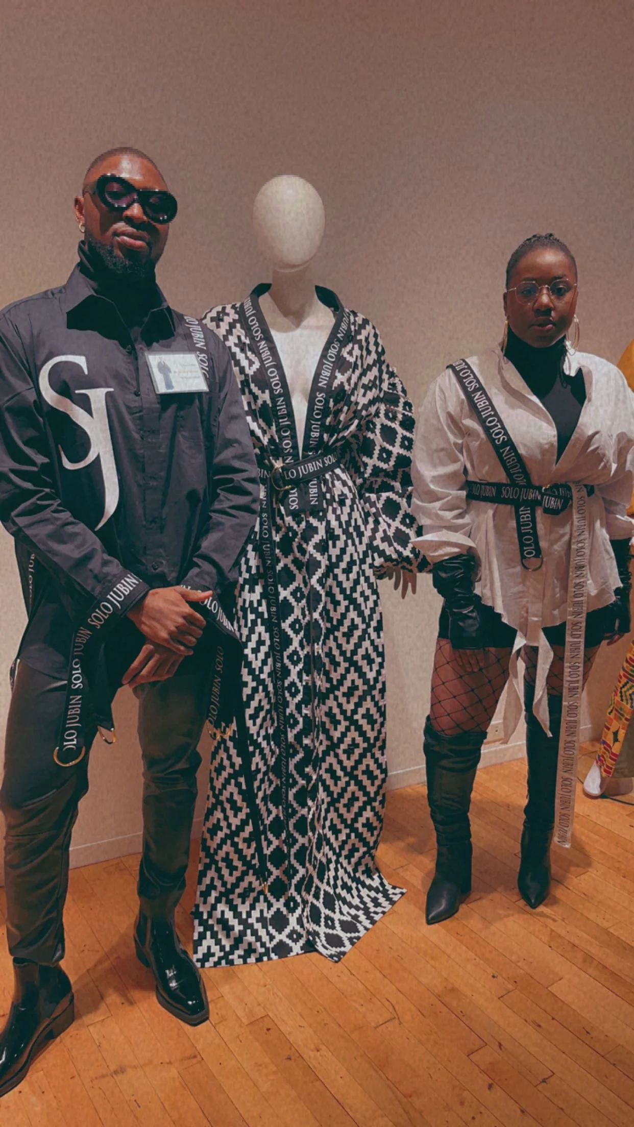 People standing next to a mannequin dressed in a geometric patterned outfit. They are wearing stylish clothing and accessories, with a neutral-colored wall and wooden floor in the background.