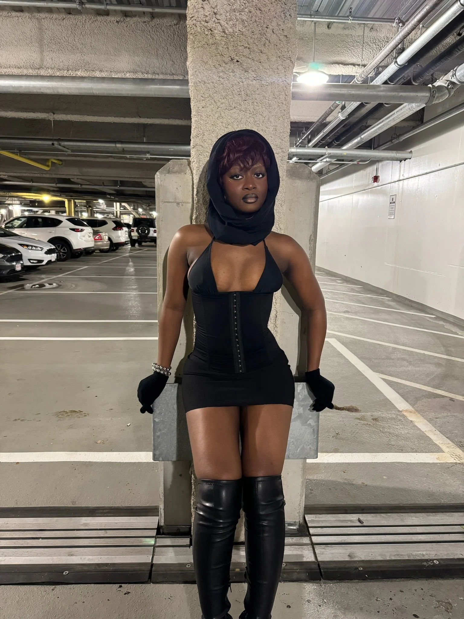 A woman dressed in black with a hooded scarf, corset, miniskirt, and thigh-high boots standing in an underground parking garage.