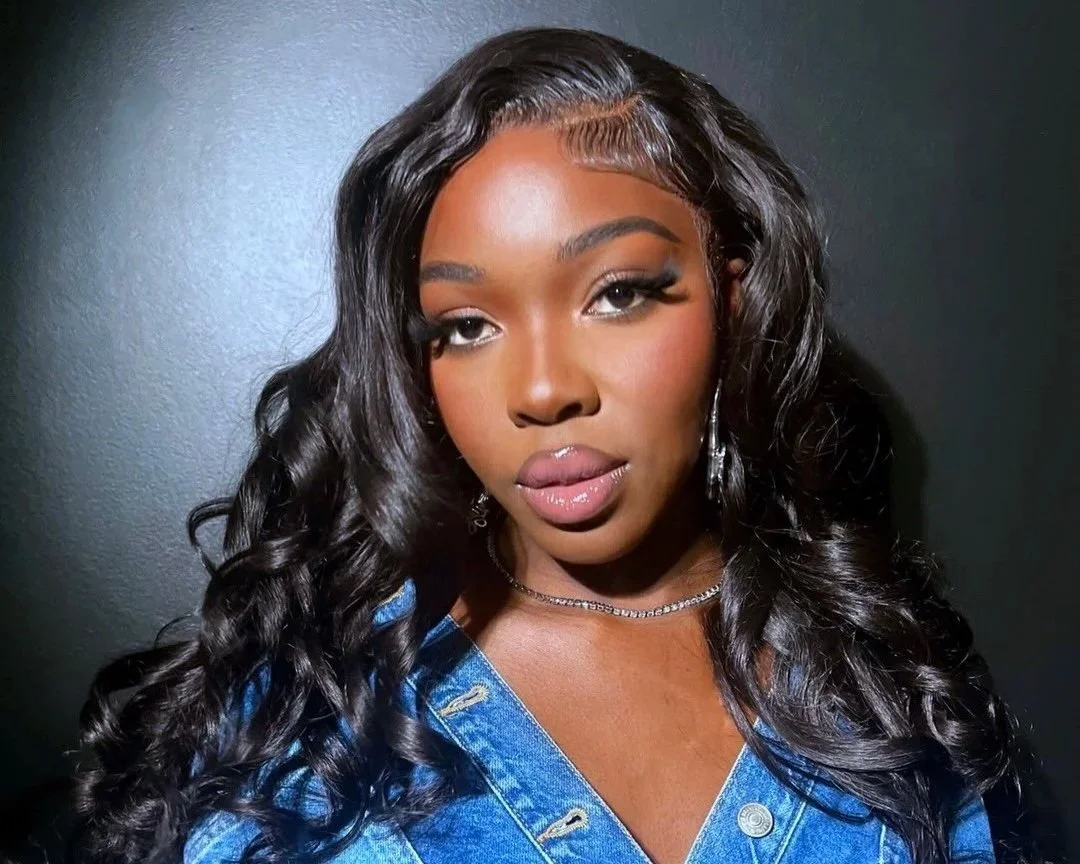 A young woman with long, wavy black hair, wearing makeup, jewelry, and a blue denim top, posing against a dark background.