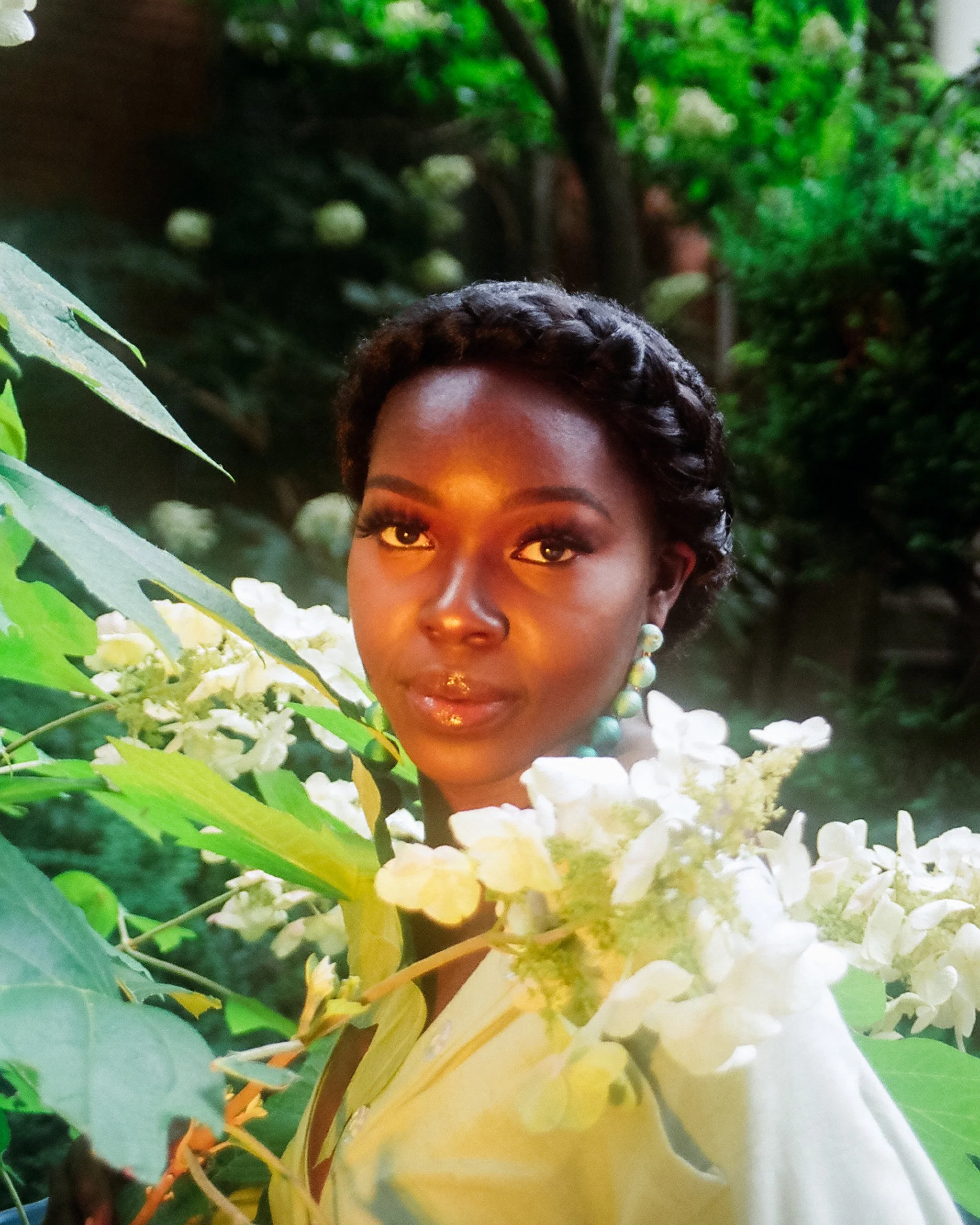 A woman with dark skin and short curly hair is surrounded by green foliage and white flowers. She is wearing turquoise beads and has a calm, confident expression.