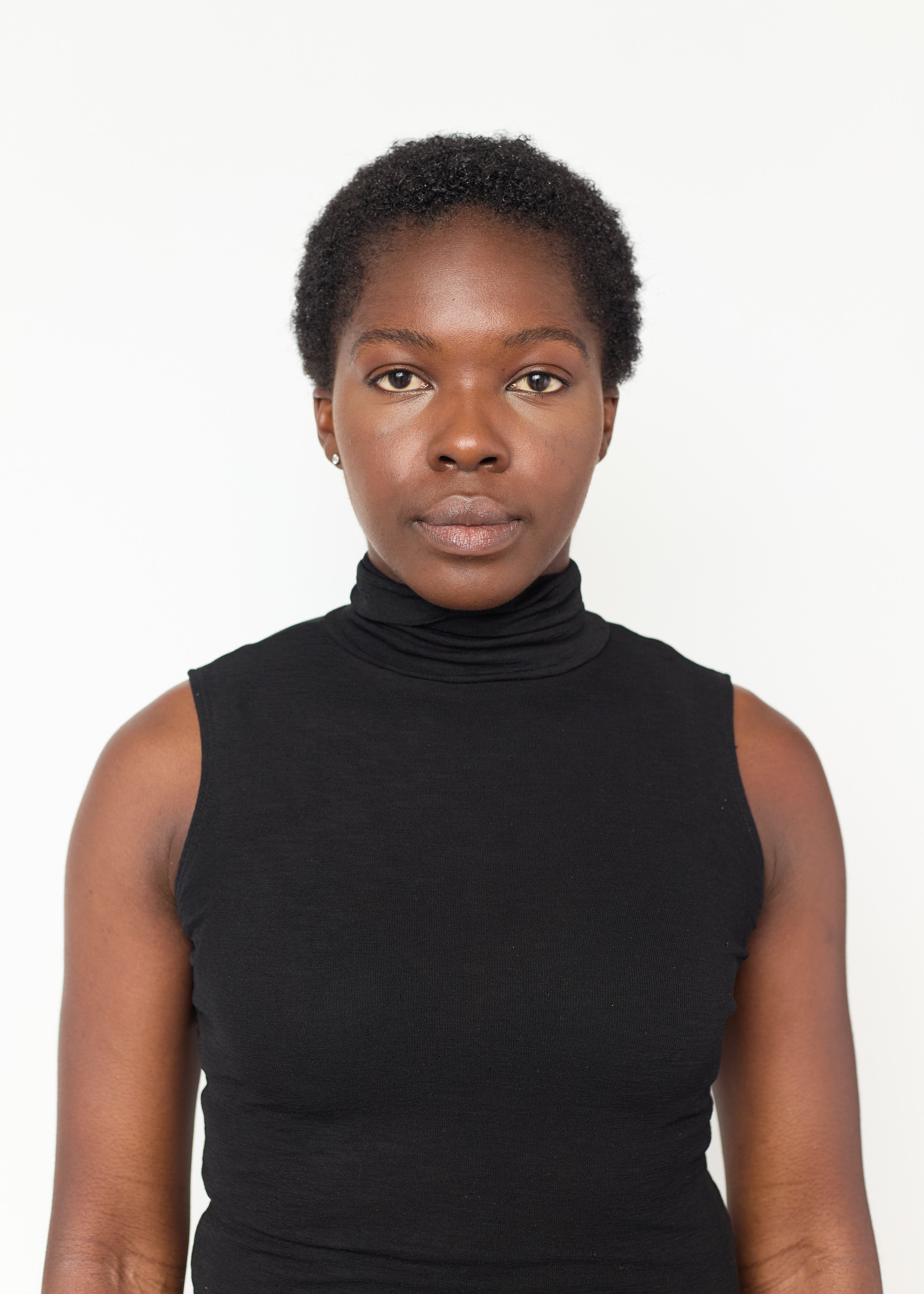 Portrait of a woman with short curly hair and brown skin, wearing a black sleeveless turtleneck top, looking directly at the camera against a plain white background.