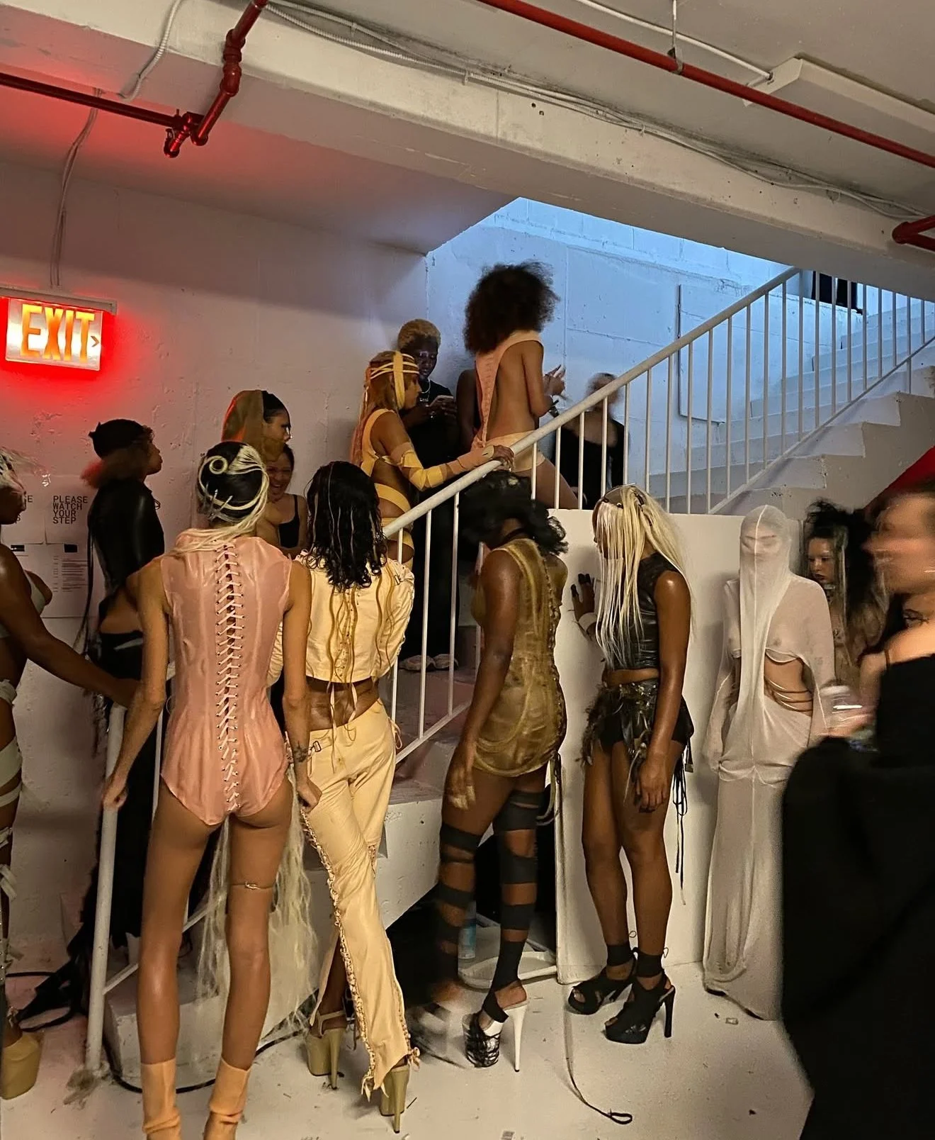 Group of people in avant-garde costumes at a fashion event or party, some standing on stairs and others on the floor, in a space with white walls, an exit sign, and industrial ceiling pipes.