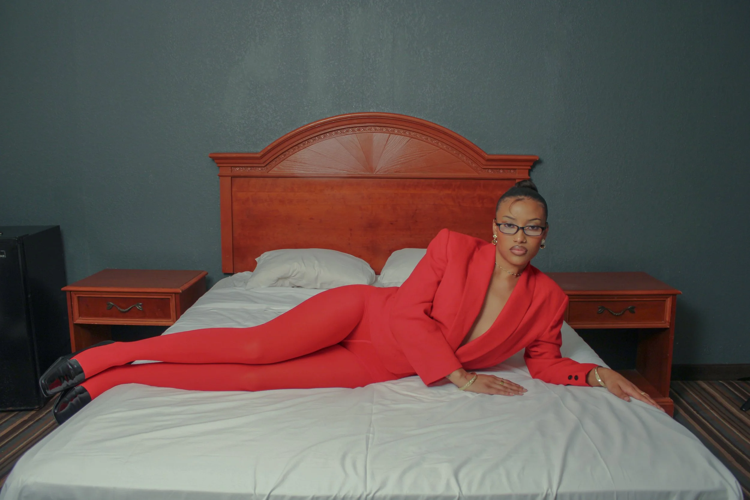 A woman in a red suit lying on a hotel bed with dark green walls and wooden headboard