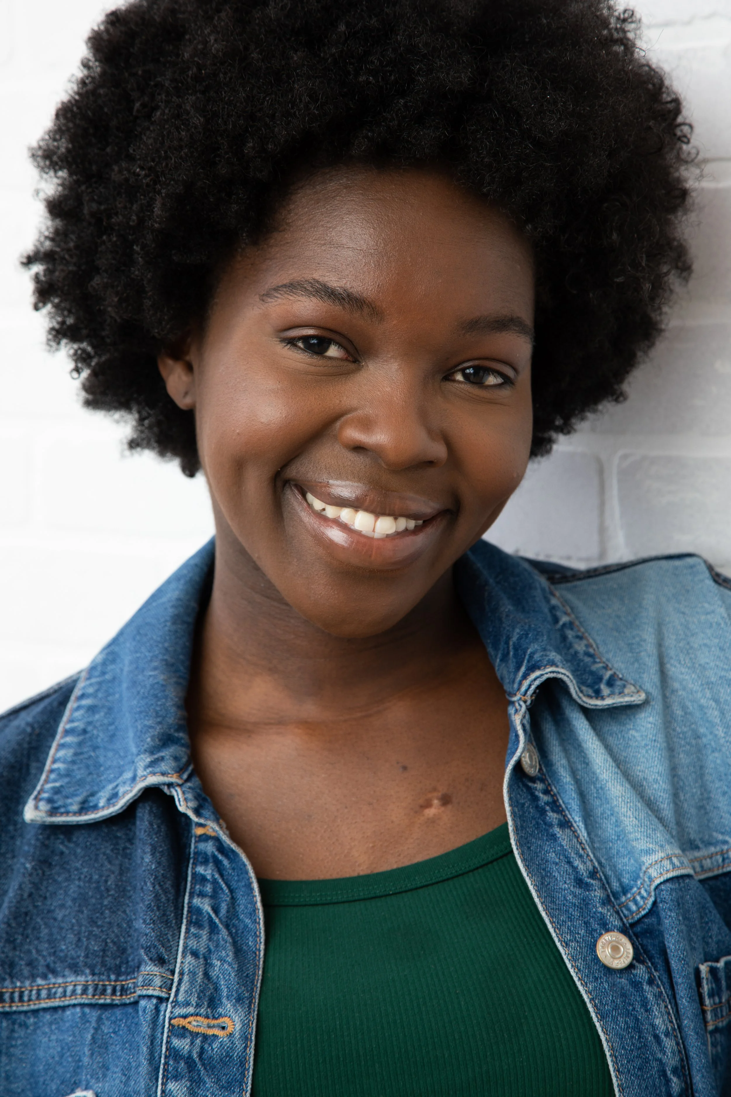 A smiling woman with short curly hair wearing a green top and denim jacket, standing in front of a white brick wall.