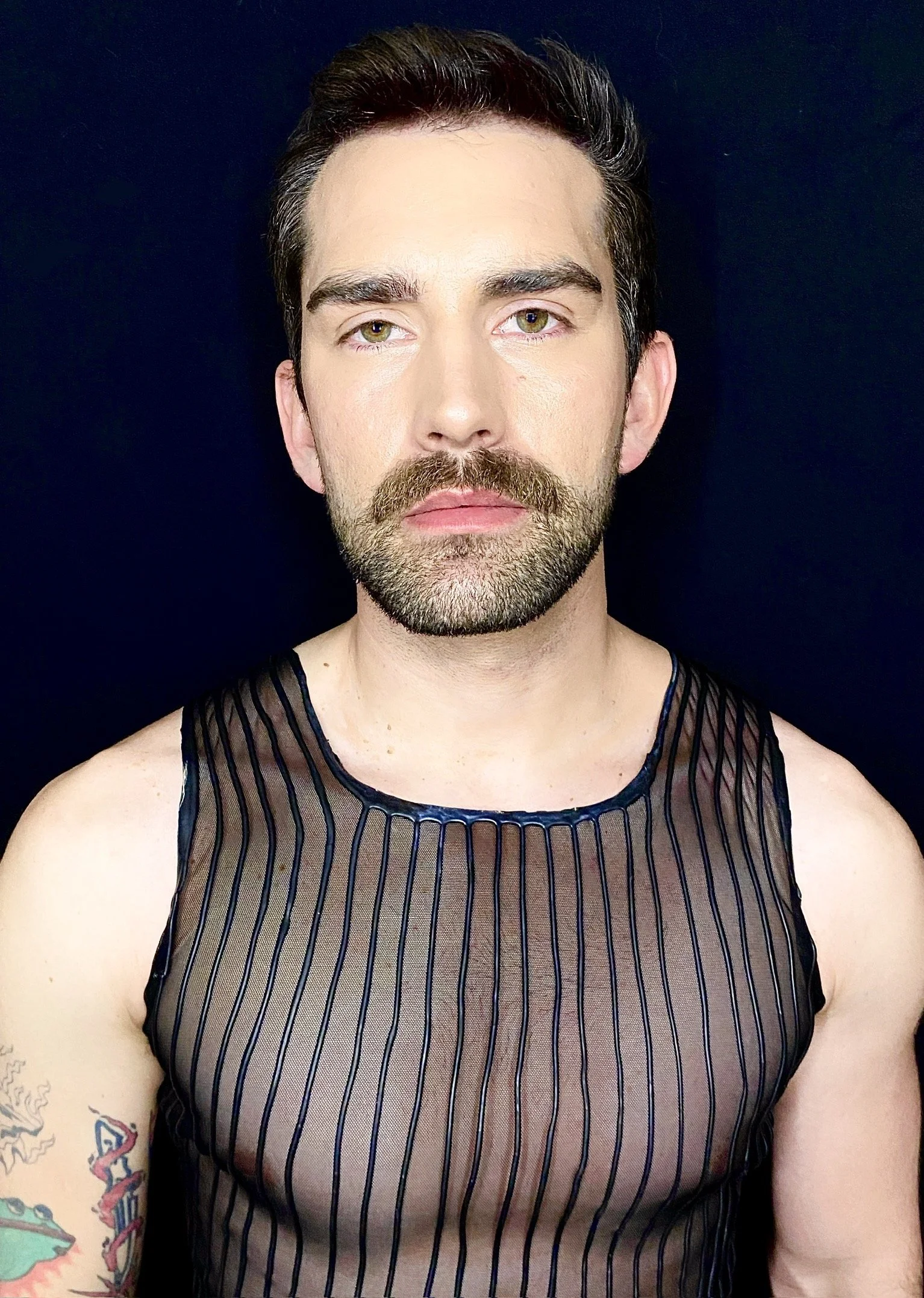 A man with brown hair, a beard, and light skin, wearing a see-through black striped tank top, standing against a dark background.
