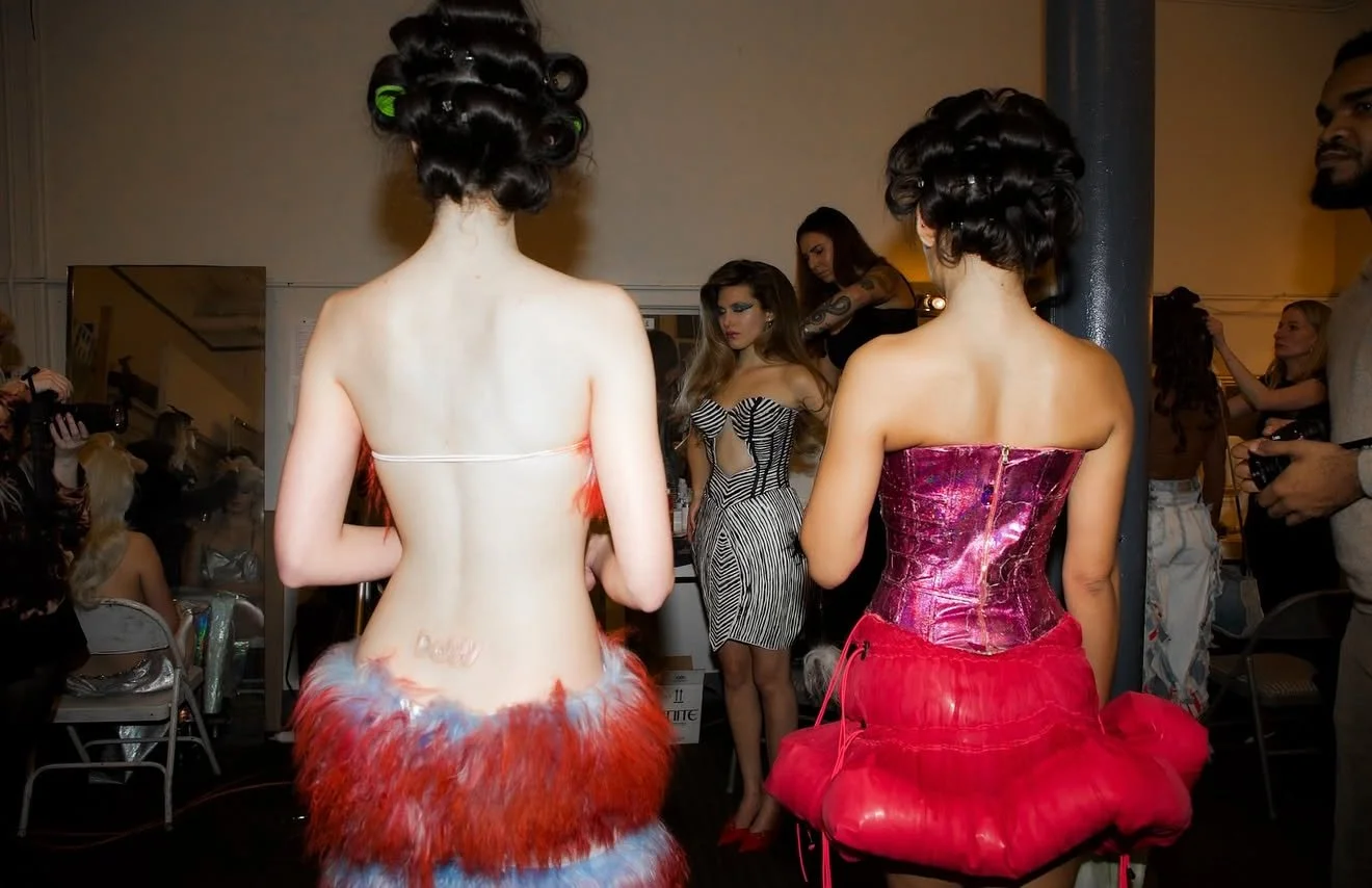 Two women with vintage-style hair in curlers and robes with puffer skirts, standing with their backs to the camera, in a room with other people and photographers.