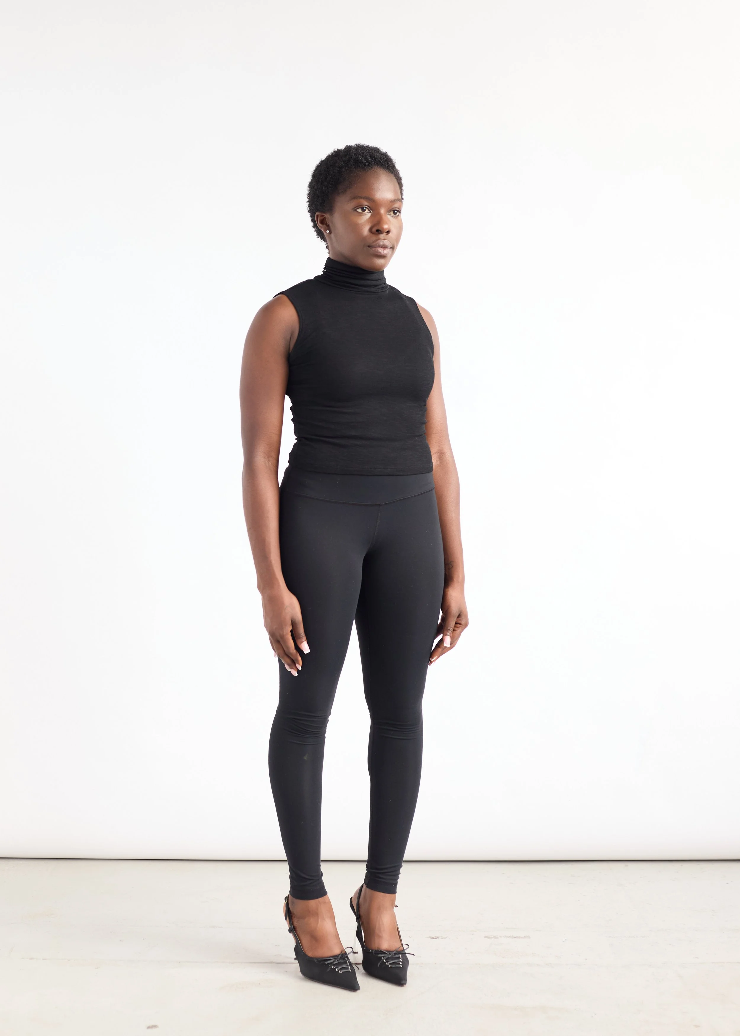 A woman in a black sleeveless turtleneck top and black leggings standing against a white background.