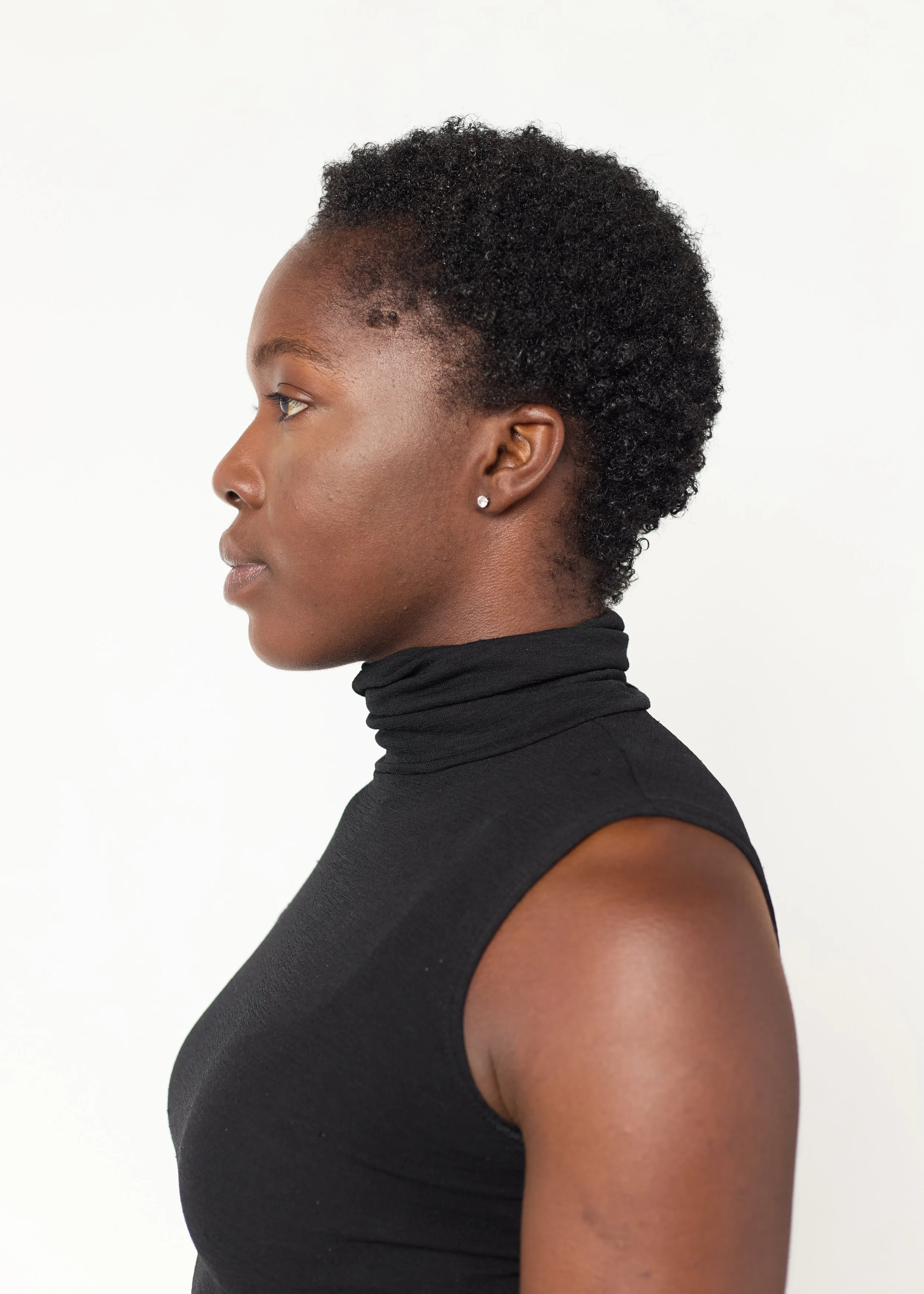 Profile of a woman with short curly hair wearing a black sleeveless turtleneck top and stud earrings, standing against a plain white background.