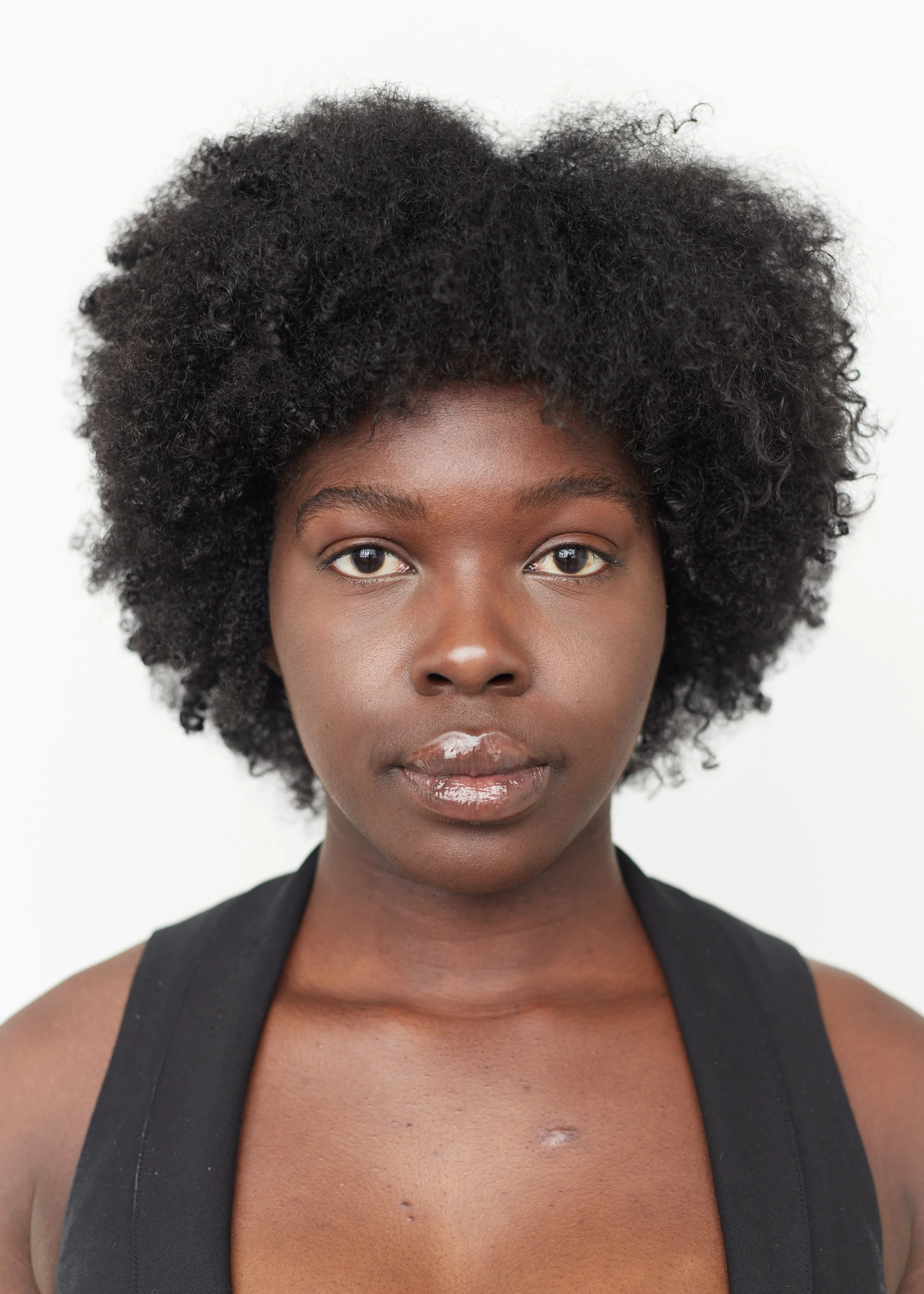 A young Black woman with a short, natural, curly afro hairstyle, wearing a sleeveless black top, facing the camera against a plain white background.
