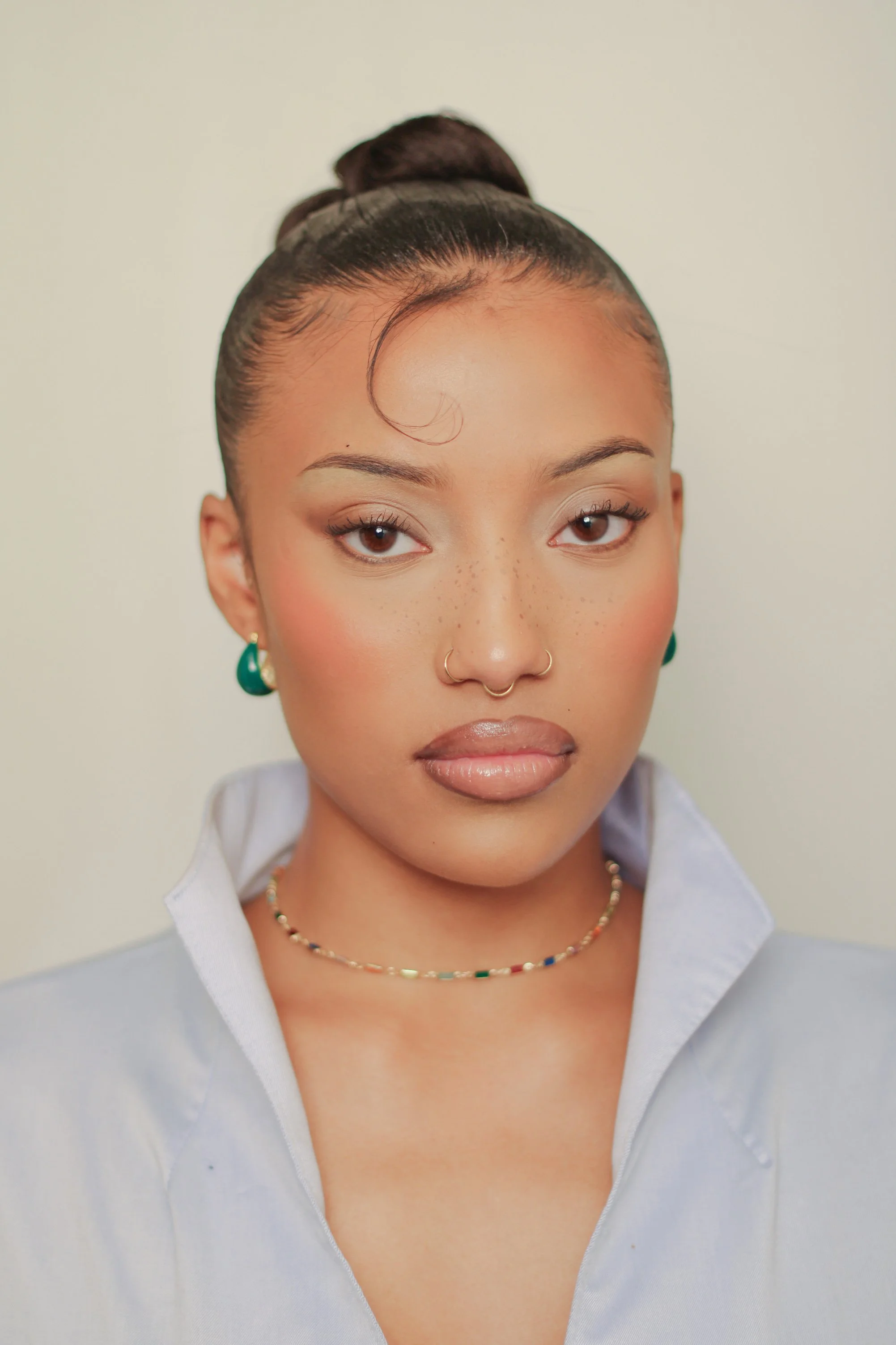 Close-up portrait of a young woman with brown hair in a neat bun, wearing a light blue collared shirt, green earrings, a multicolored beaded necklace, and gold nose rings, looking directly at the camera against a plain, light-colored background.