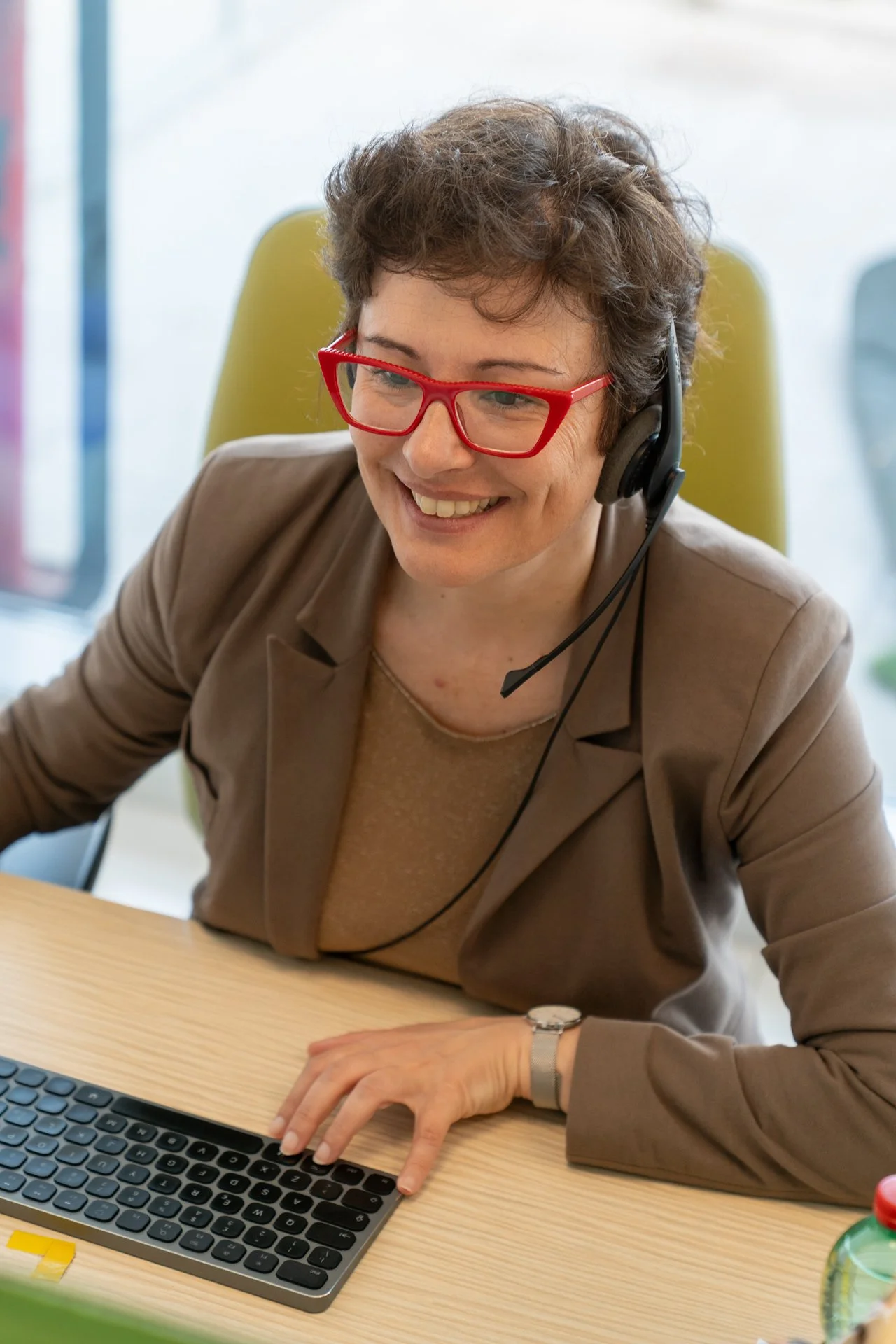 Donna sorridente con occhiali rossi, ascoltando con un auricolare, mentre lavora al computer in ufficio.