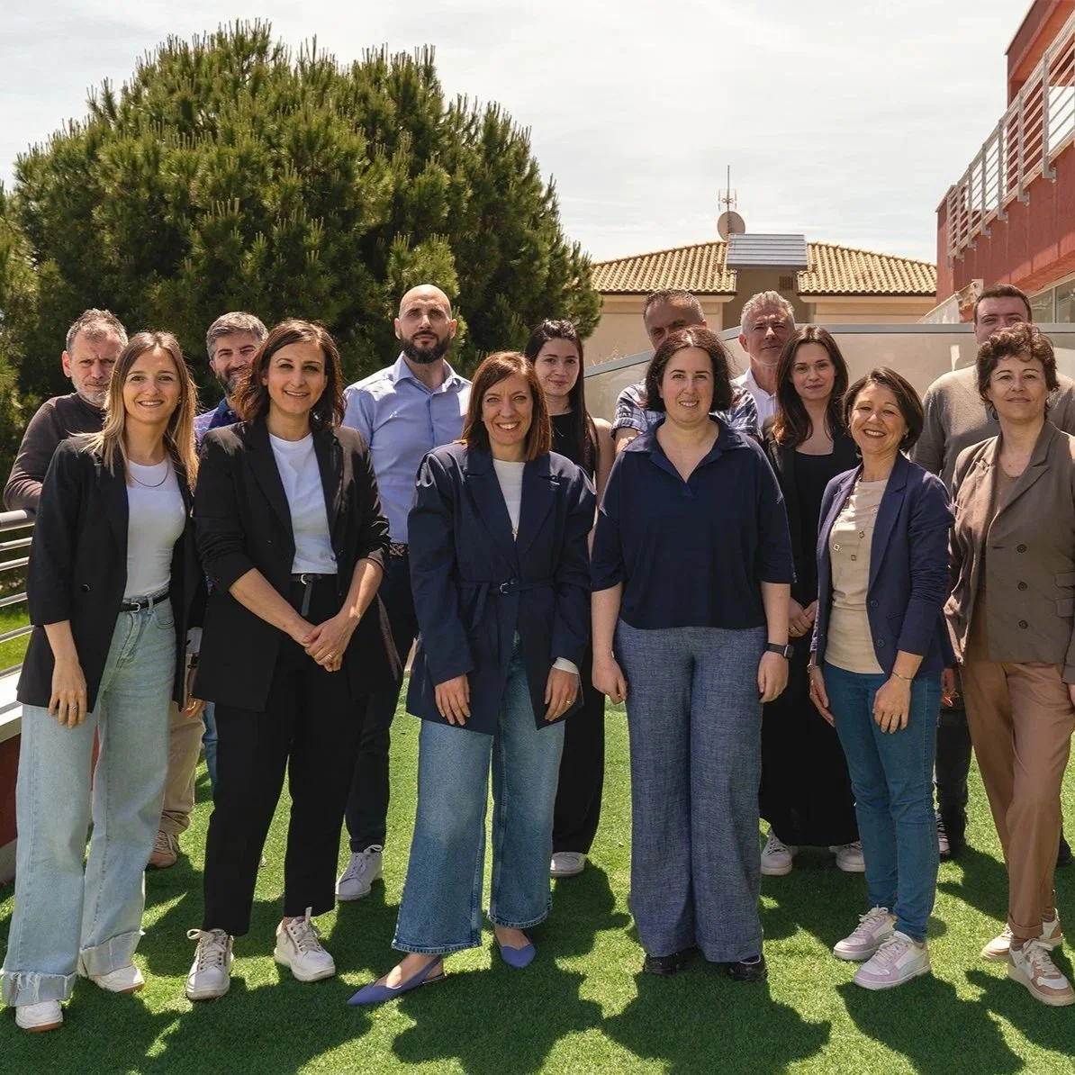 Gruppo di persone sorridenti posano per una fotografia all'aperto, alcuni indossano abiti business casual, in un ambiente con prato verde e alberi sullo sfondo.