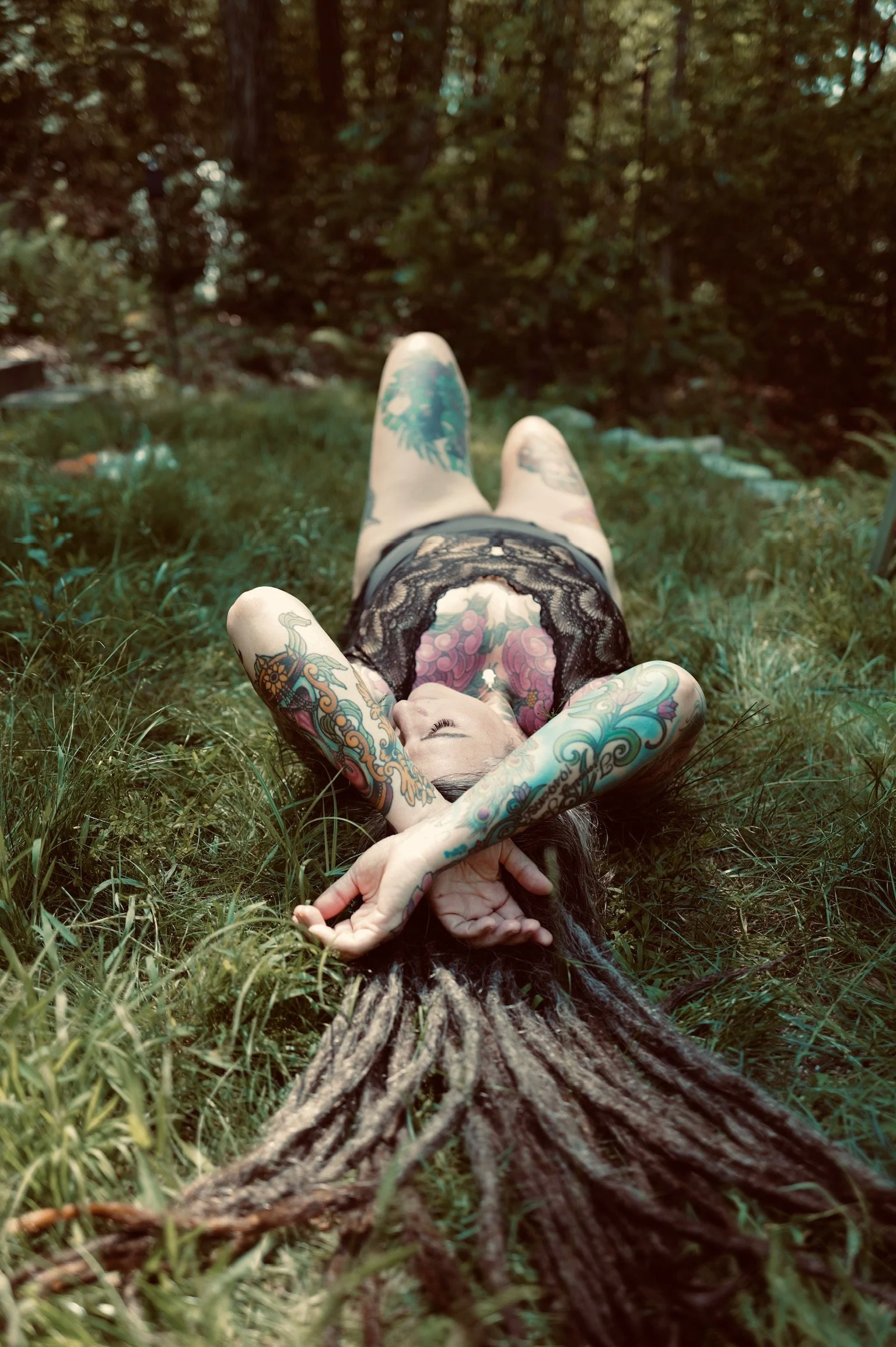 A woman with tattoos, lying on her back on grass in a forest, with legs raised and arms behind her head, wearing black lace clothing. Stacy Crouse Siddhanimomma of Siddhani Wellness
