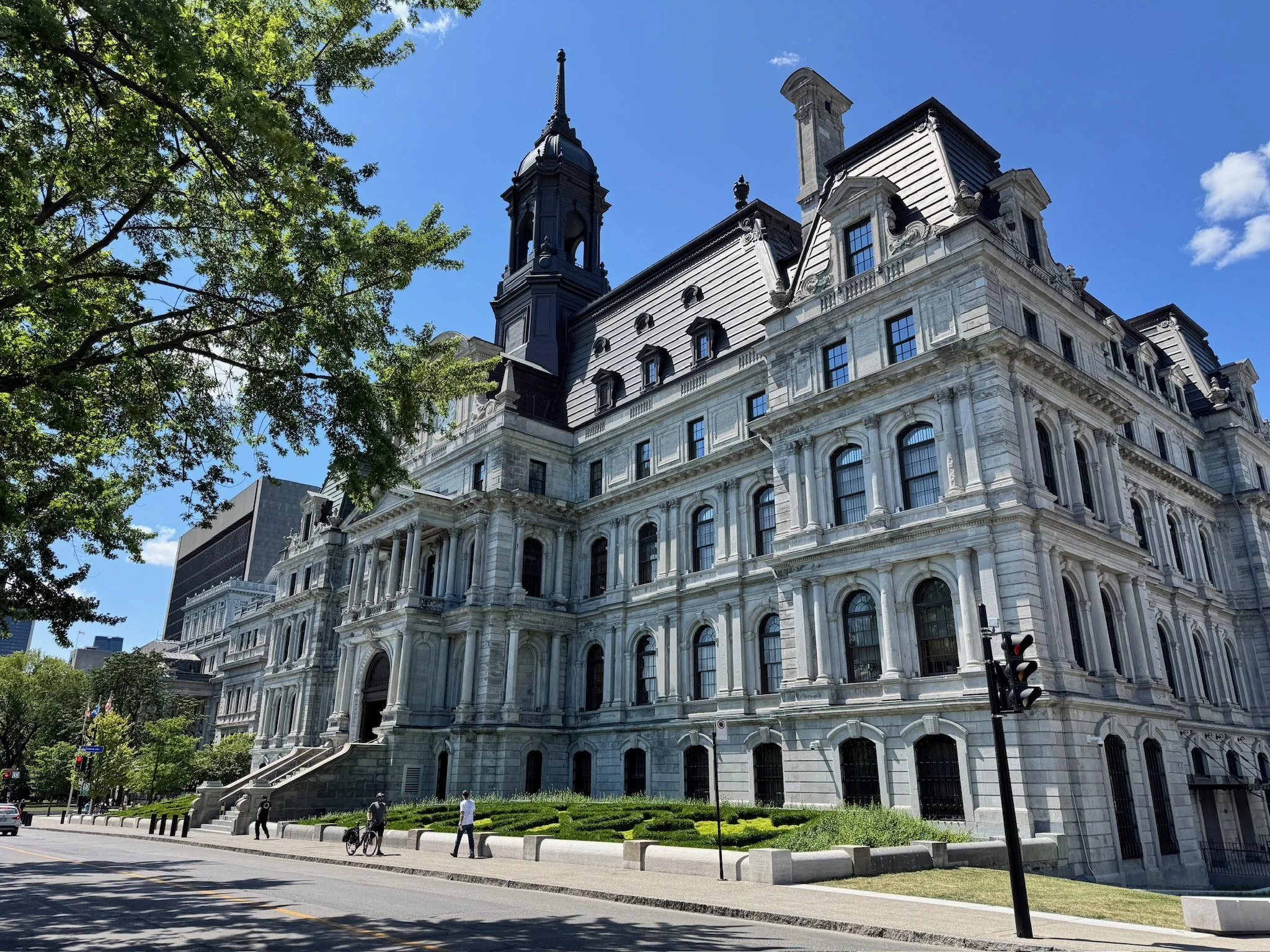 A large historic government building in Old Montreal Canada with elaborate architecture, white stone facade, arched windows, a dome with a clock, and a black turret against a blue sky.
