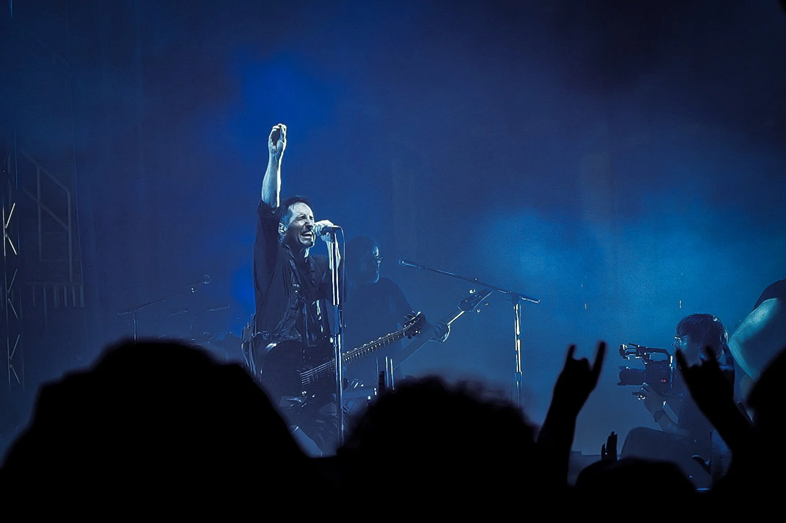 Trent Reznor of Nine Inch Nails on stage at TD Garden in Boston Massachusetts singing into microphone, playing guitar, with their right arm raised, in a dark blue-lit concert setting with an audience and a camera person in the foreground.