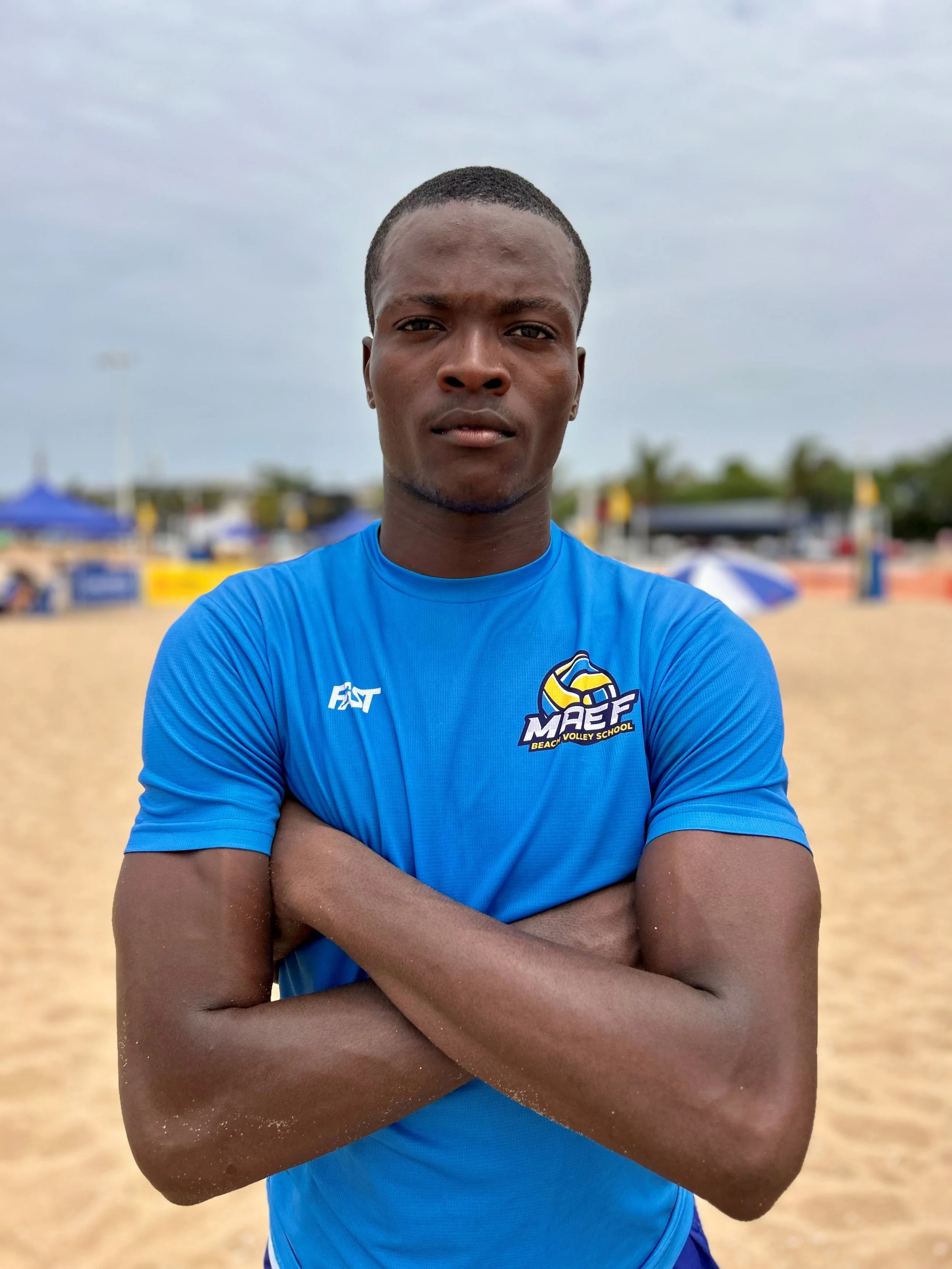 Young male athlete standing outdoors on a sandy track, wearing a white sports jersey with various logos and the number 1, under a shaded canopy with a sports field and buildings in the background.