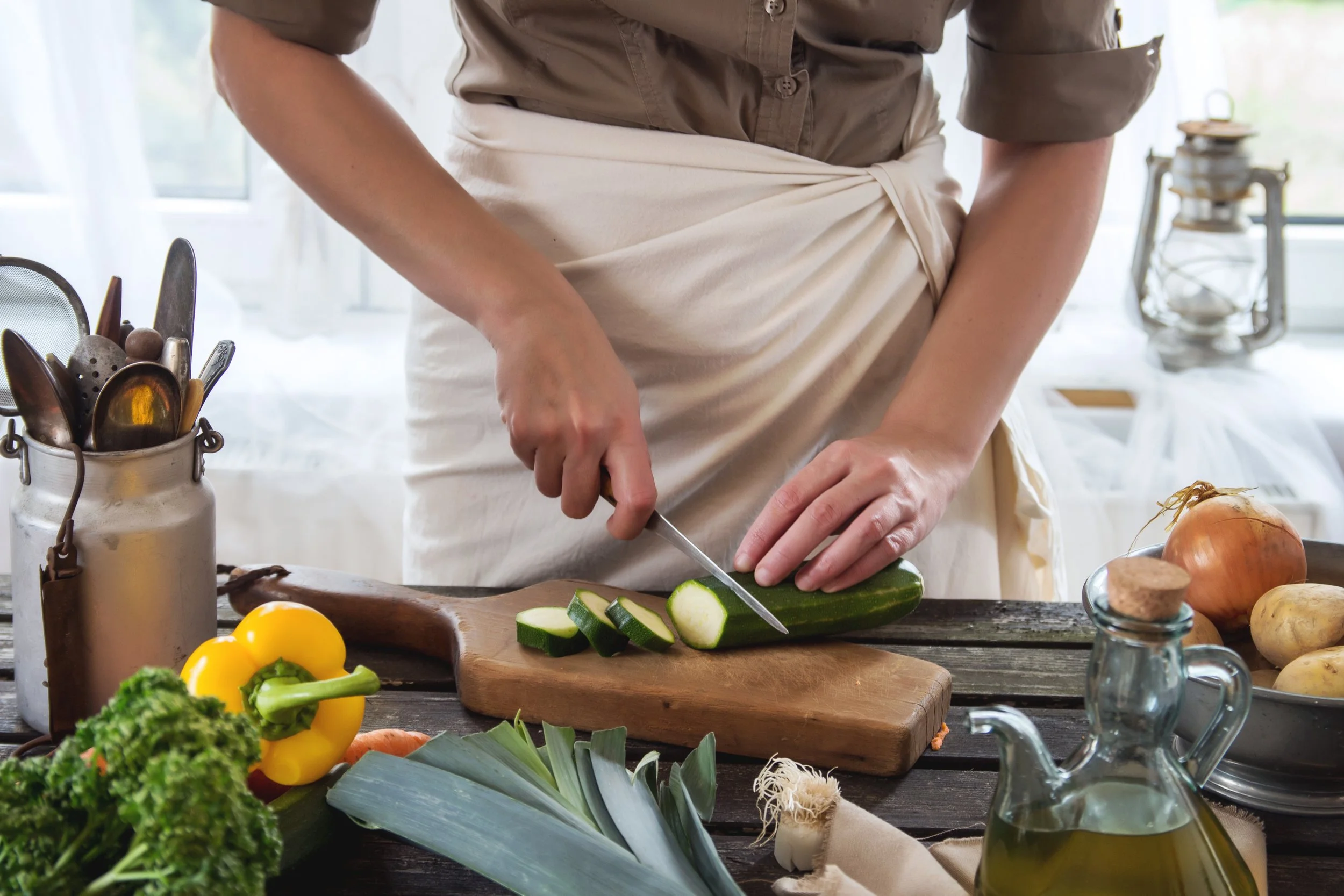 A person chopping cucumbers on a wooden cutting board surrounded by various vegetables and kitchen utensils in a well-lit kitchen.