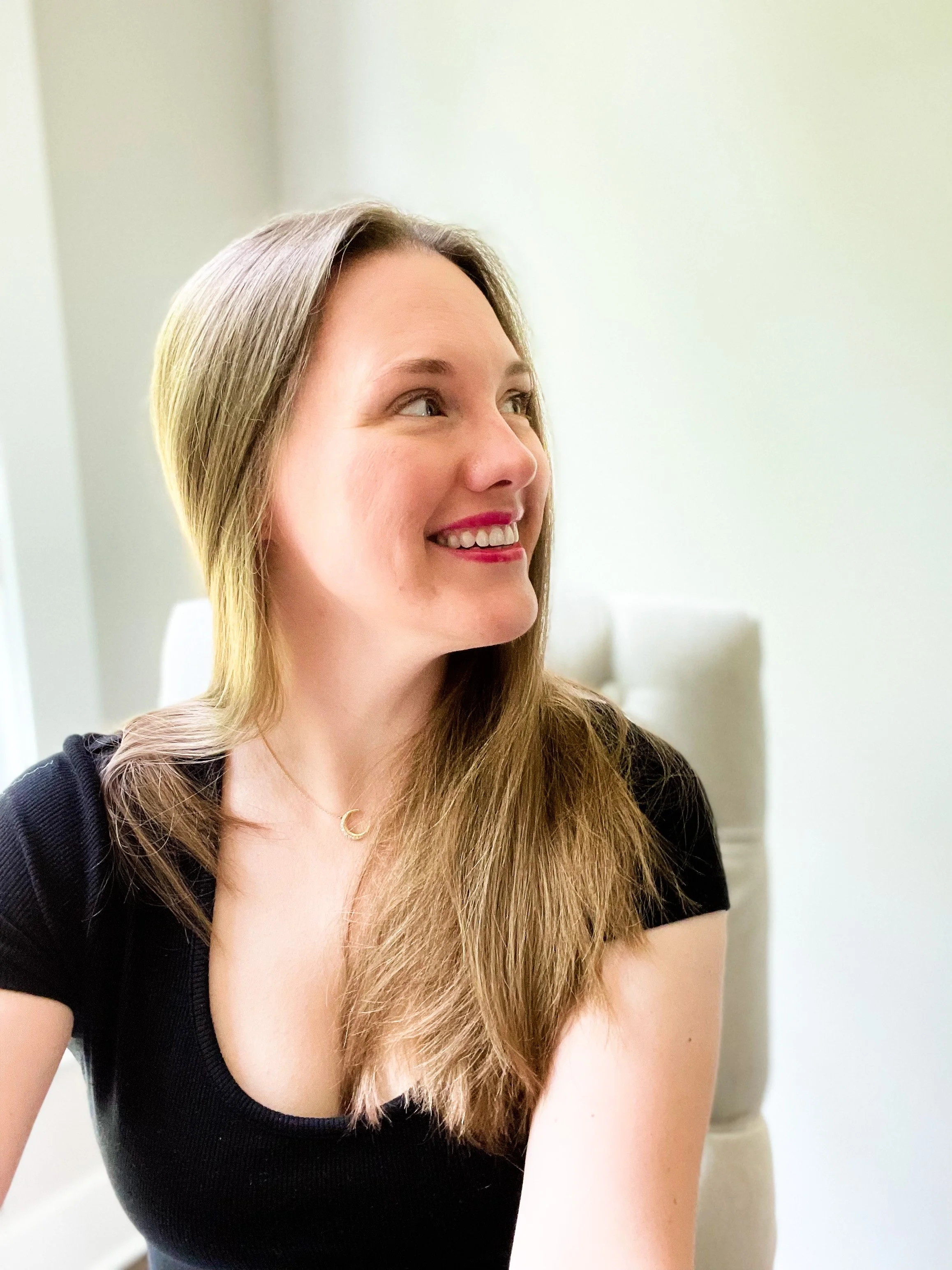 Close-up of a young woman with long blonde hair, smiling and looking to the side, wearing a black top and a delicate gold necklace with a small 'C' pendant, in a well-lit indoor setting.