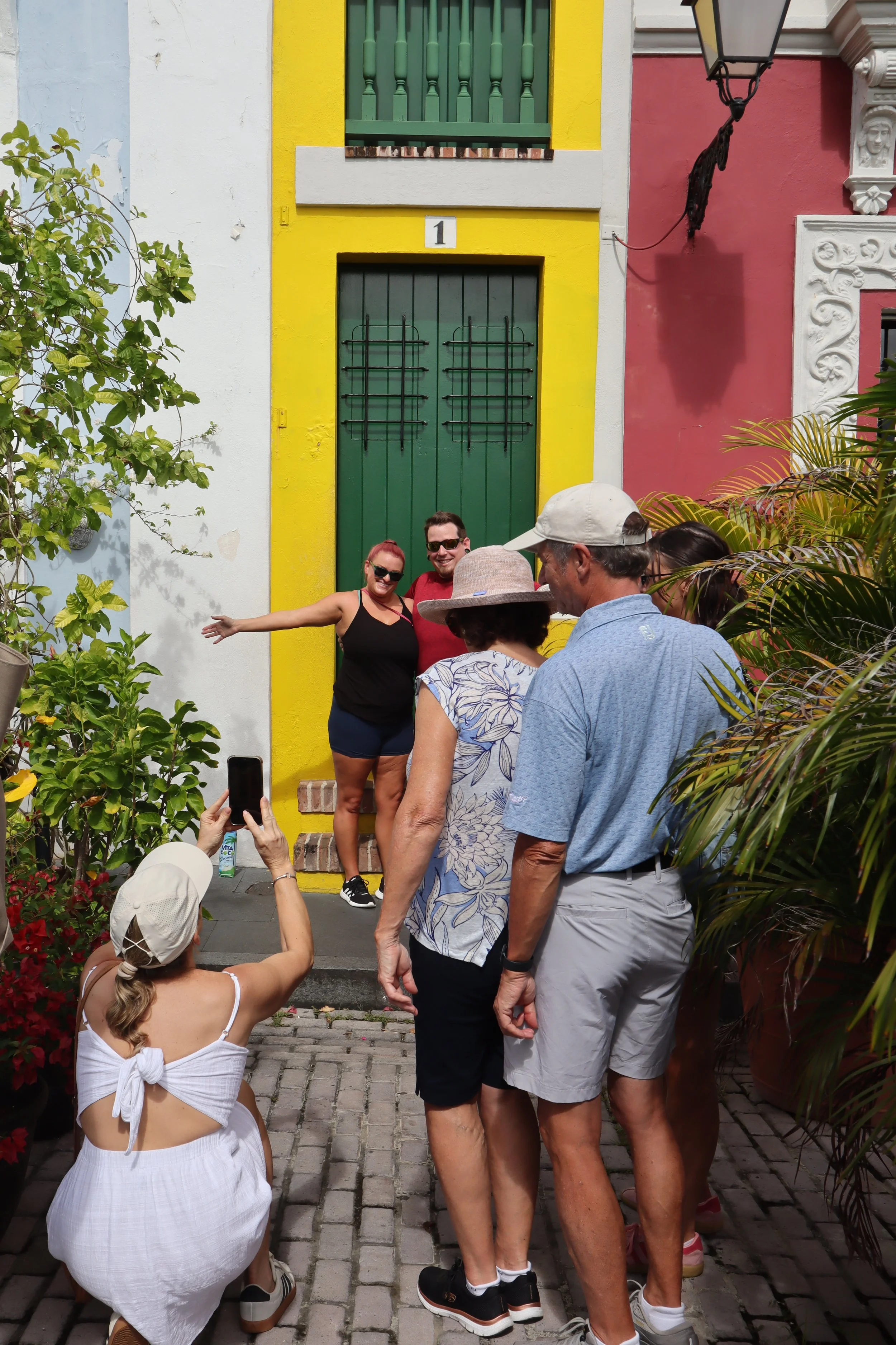 A group of people taking a photo in front of a colorful building with yellow, white, and pink walls. Two people are posing in front of a green door while others watch and take pictures. The scene appears to be lively and cheerful.