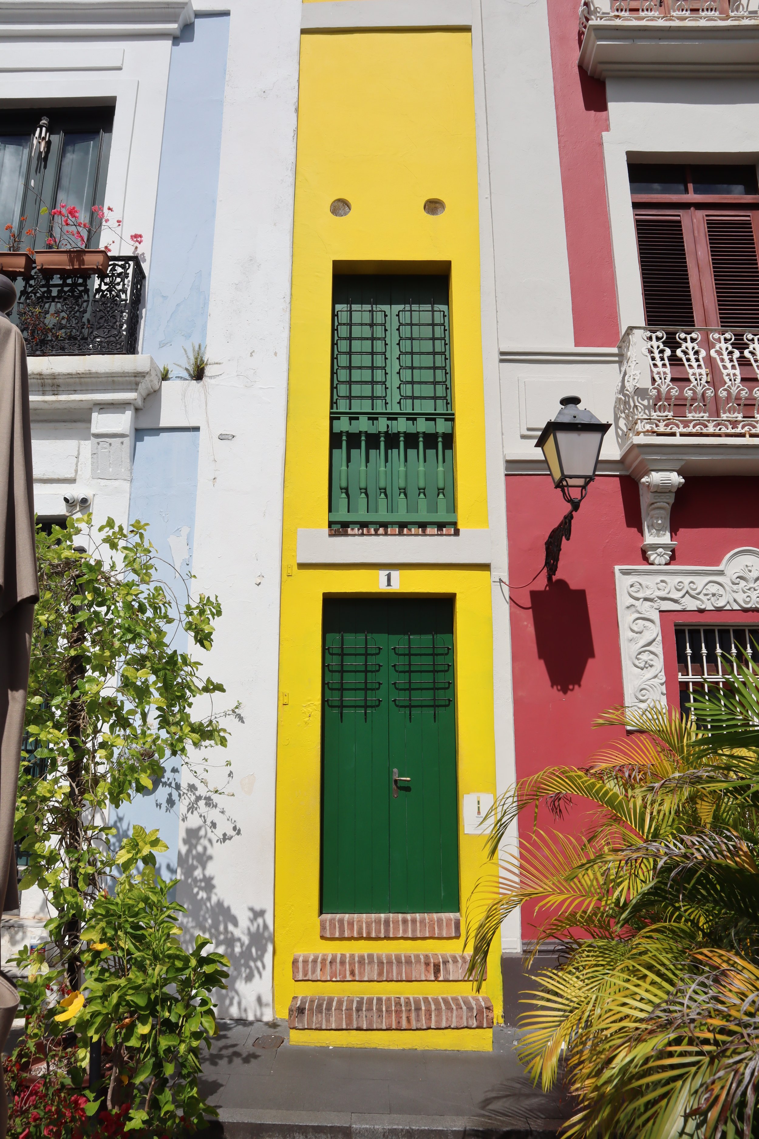 Colorful building with a yellow front and green door and window shutters, flanked by white and red buildings, with plants and a street lamp.