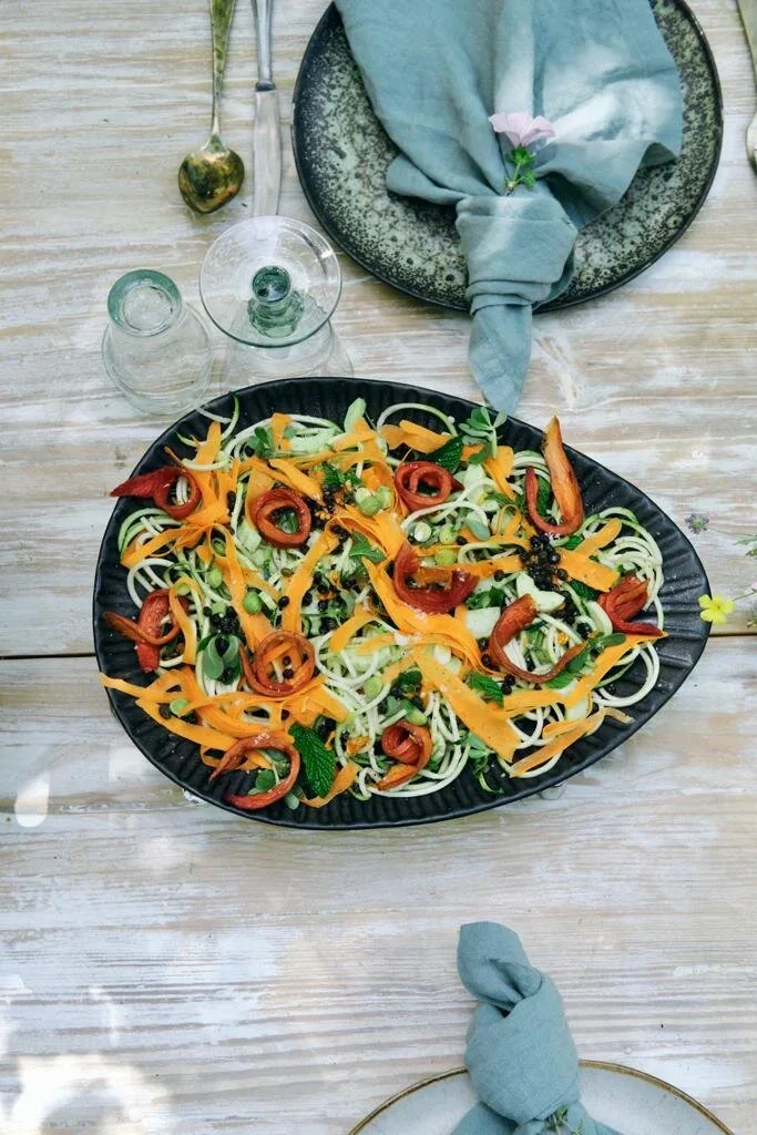 A black oval plate with a salad that includes spiralized zucchini, red bell pepper rings, shredded carrots, chopped green onions, and black sesame seeds, on a wooden table with a teal cloth napkin, a small bouquet of pink flowers, and two glasses of 