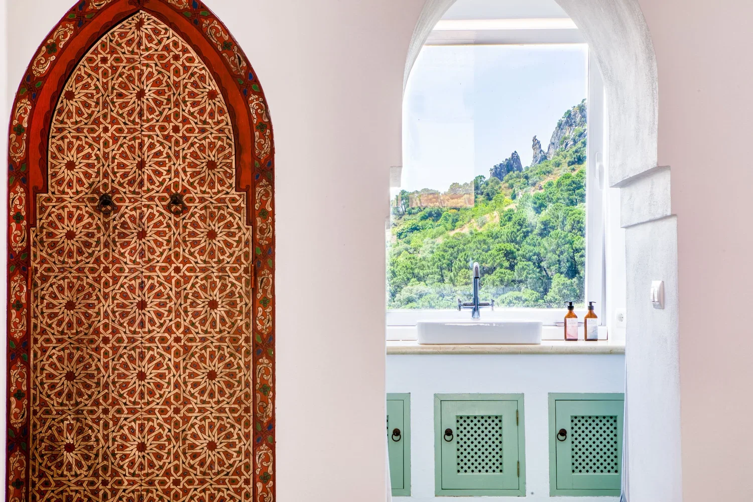 Interior of a room with a white wall, a decorative arched niche with intricate geometric patterns, and a window with a scenic view of green hills and rocky formations. The window has a white frame and a countertop with two soap dispensers.
