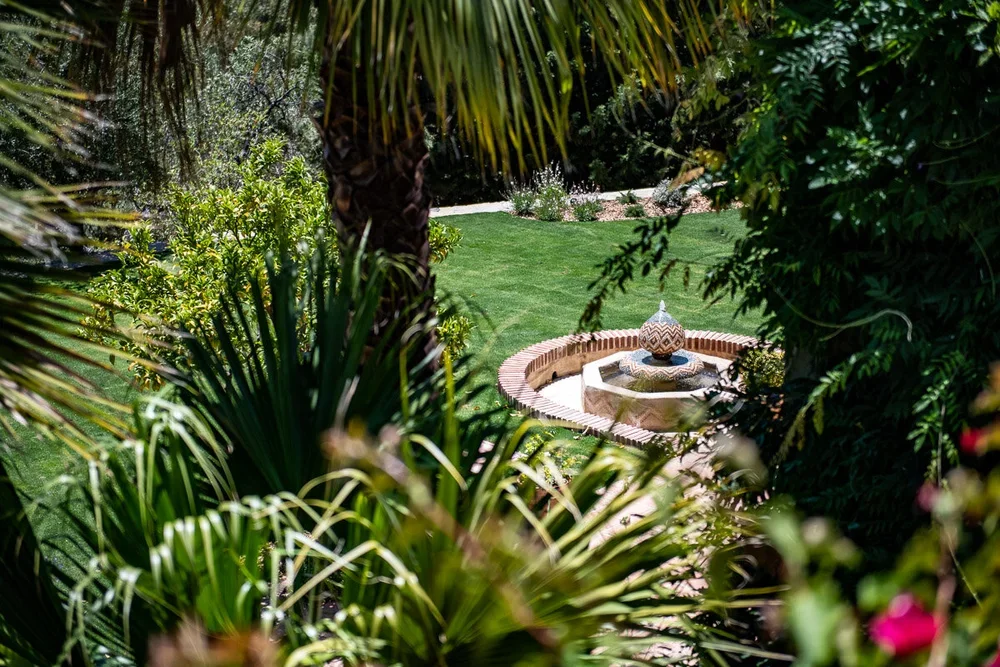 Lush garden with palm trees, bushes, and a decorative fountain with a ceramic ornament on top, surrounded by a brick border.