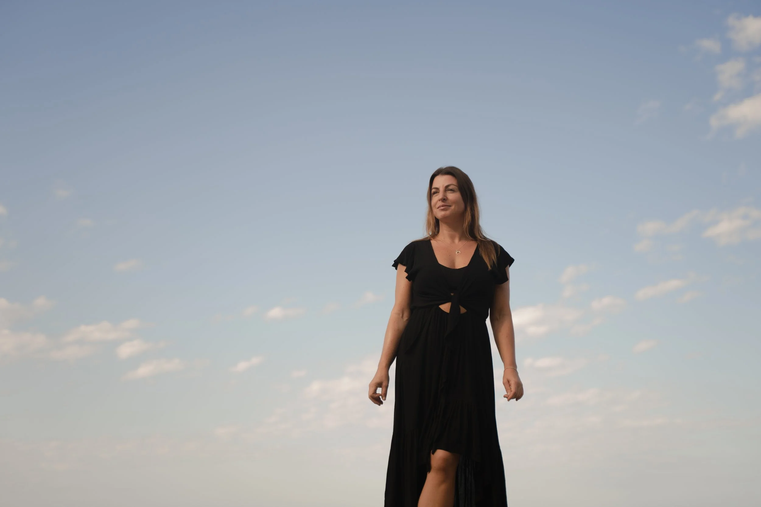 Woman in a black dress walking outdoors under a blue sky with scattered clouds.