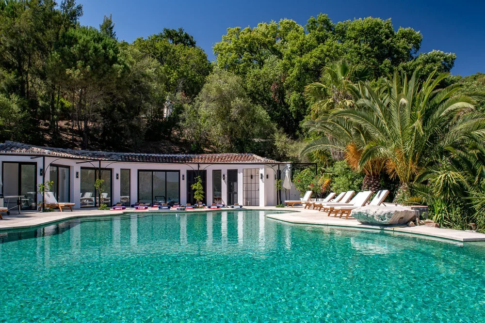 A luxurious outdoor swimming pool surrounded by lounge chairs and lush green trees under a clear blue sky.