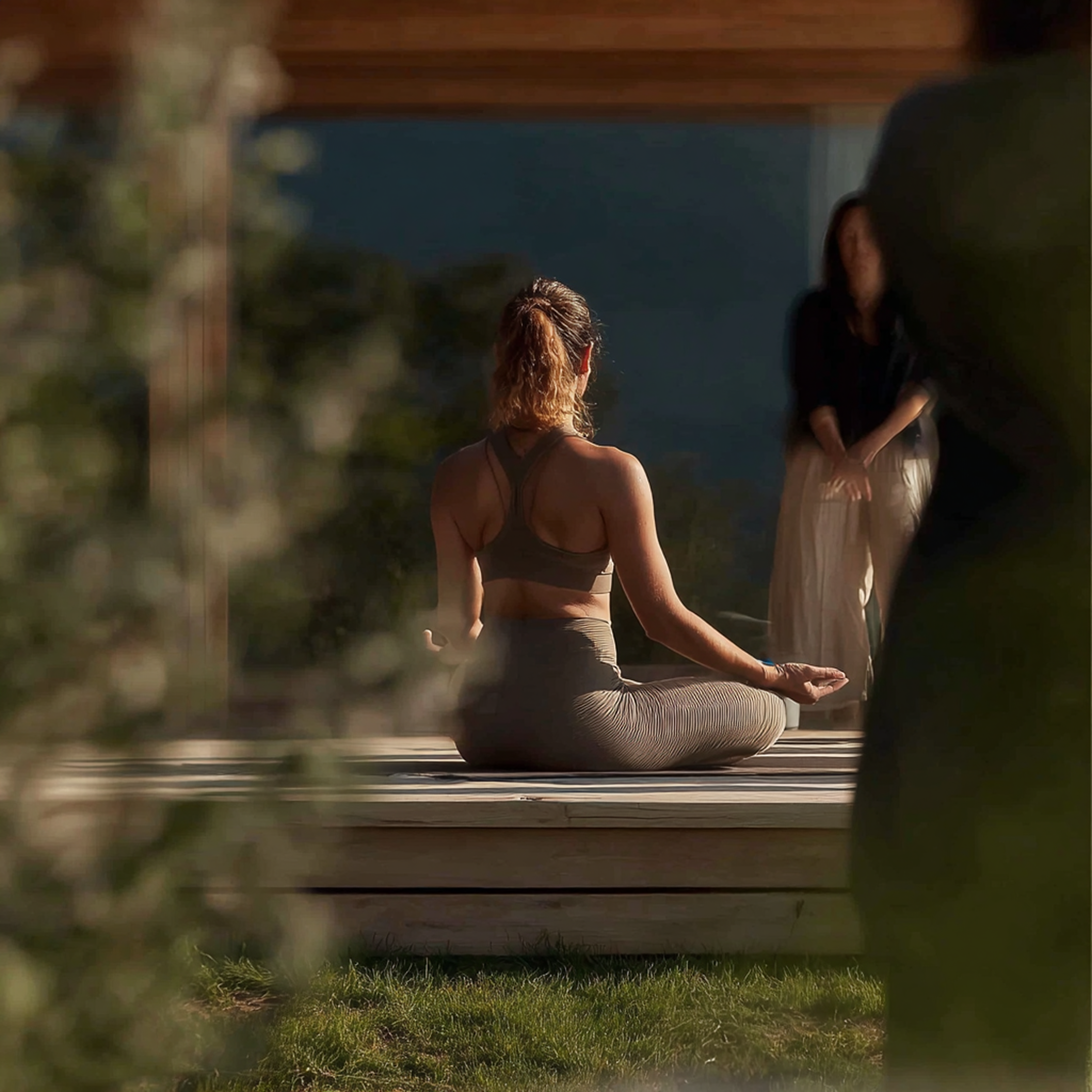A woman practicing yoga outdoors in a seated meditation pose on a wooden platform, with another person standing nearby and a third person in the background, during the evening or early morning.