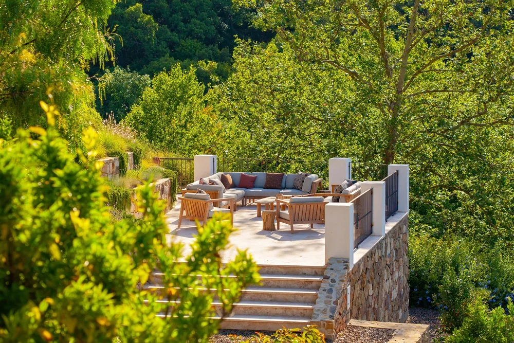 Outdoor patio with seating area, including a sectional sofa, wooden chairs, and tables, surrounded by lush green trees and plants.