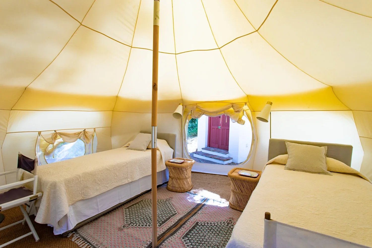 Interior of a bell tent with two single beds, small wicker side tables, a patterned rug, and an open entrance showing steps and a red door outside