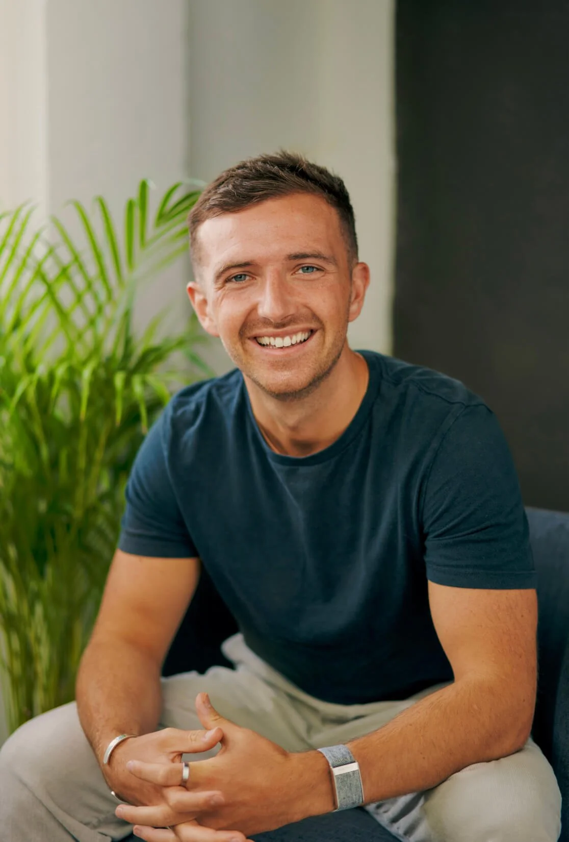A smiling man with short brown hair and blue eyes, wearing a navy blue t-shirt, sitting indoors with a green plant in the background.