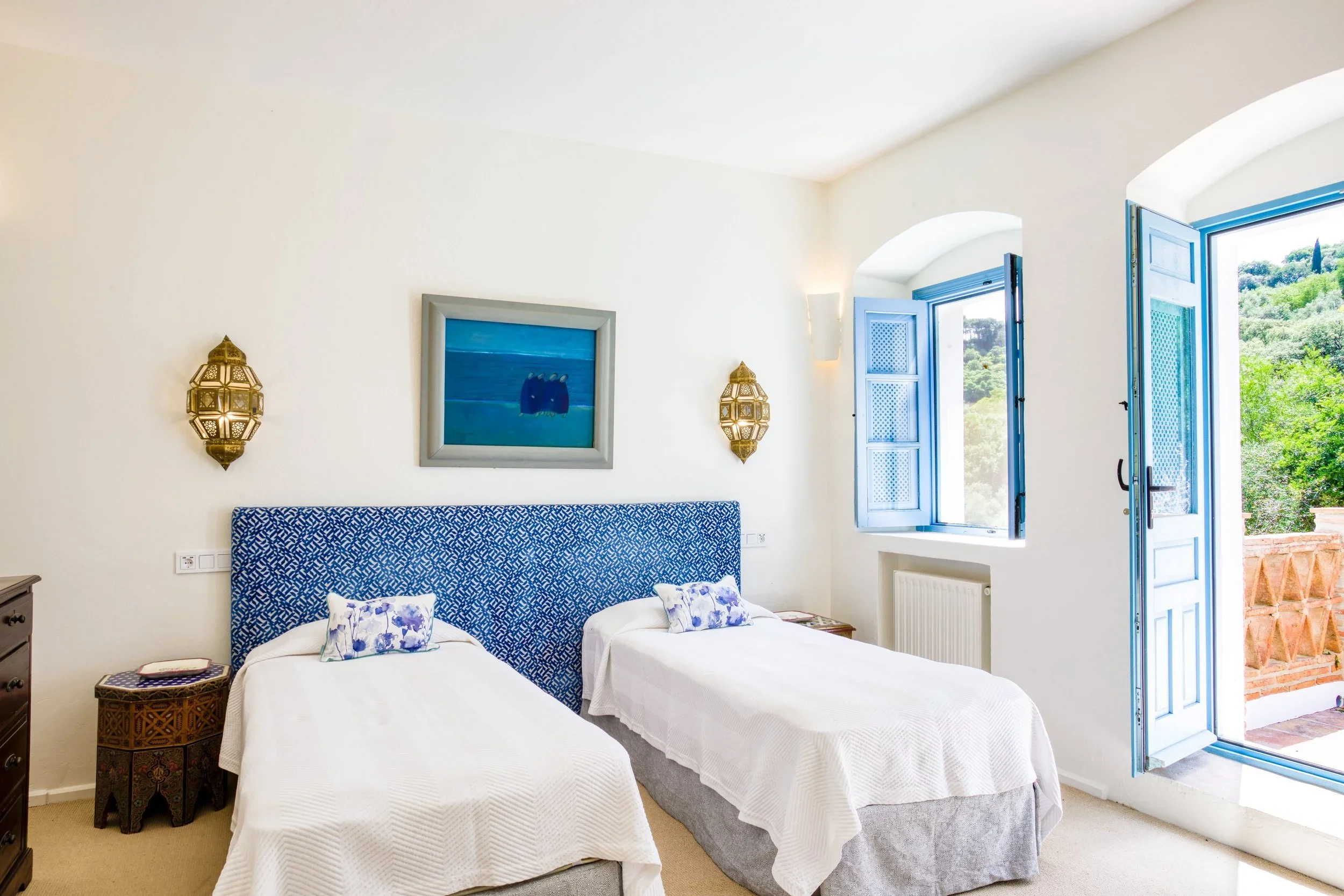 A bright bedroom with two twin beds dressed in white linens, a blue patterned headboard, and decorative pillows. There are two ornate wall lamps, a framed ocean-themed painting, and open blue shutters on a window revealing a view of lush greenery outside.
