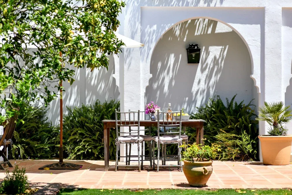Outdoor patio with a wooden dining table, four chairs, potted plants, and a white wall with arched architectural features and shadows of palm leaves.