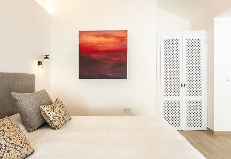A bedroom with a bed, several pillows, wall-mounted light, and colorful abstract painting on the wall, with a white closet door on the right side.