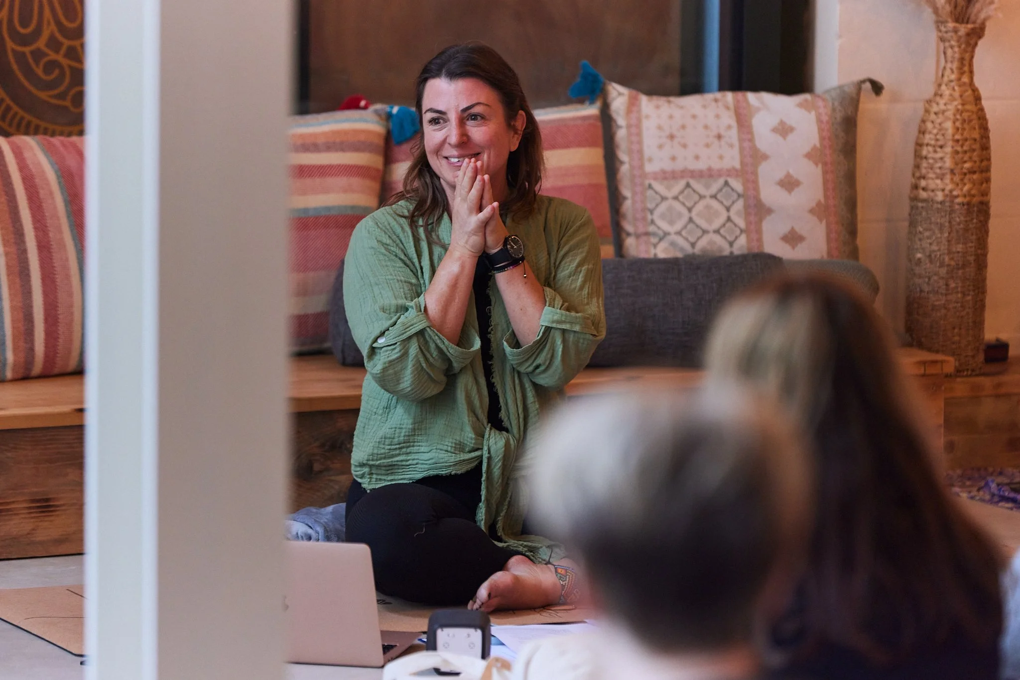 Woman smiling and gesturing with hands during a virtual meeting or class, sitting on a floor with a laptop, in a cozy room with pillows and decorative items.